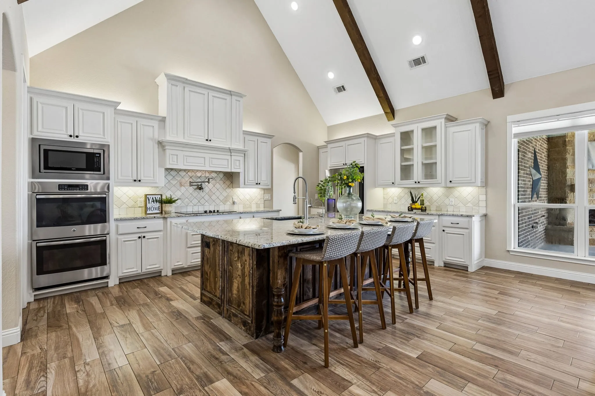 Vaulted ceiling with exposed beams, wood-finish flooring, white cabinetry with under-cabinet lighting, stainless steel double ovens and microwave, and a large center island with a wood-clad base