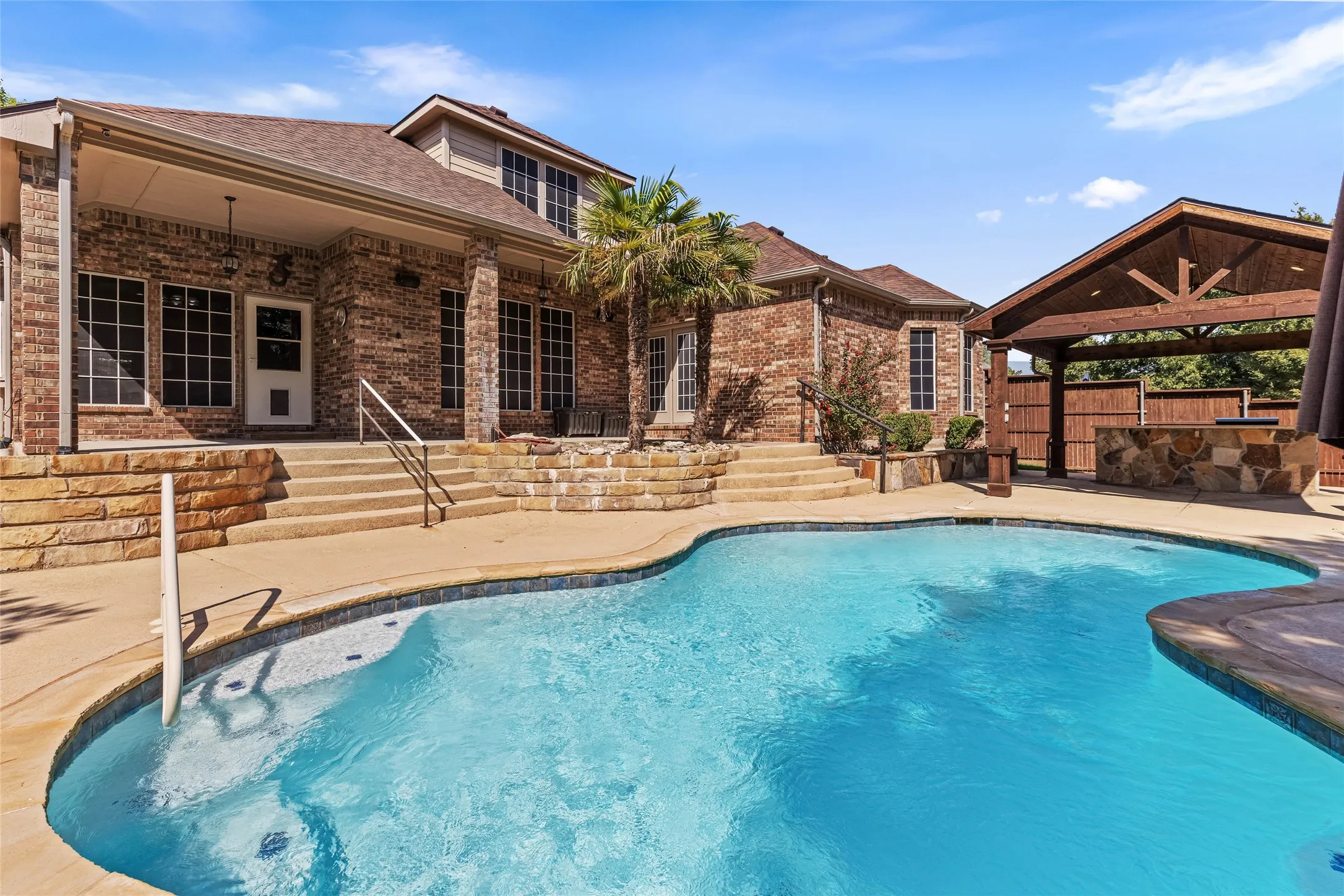 Brick exterior with a covered patio, freeform swimming pool, and stone-clad outdoor kitchen under a pergola