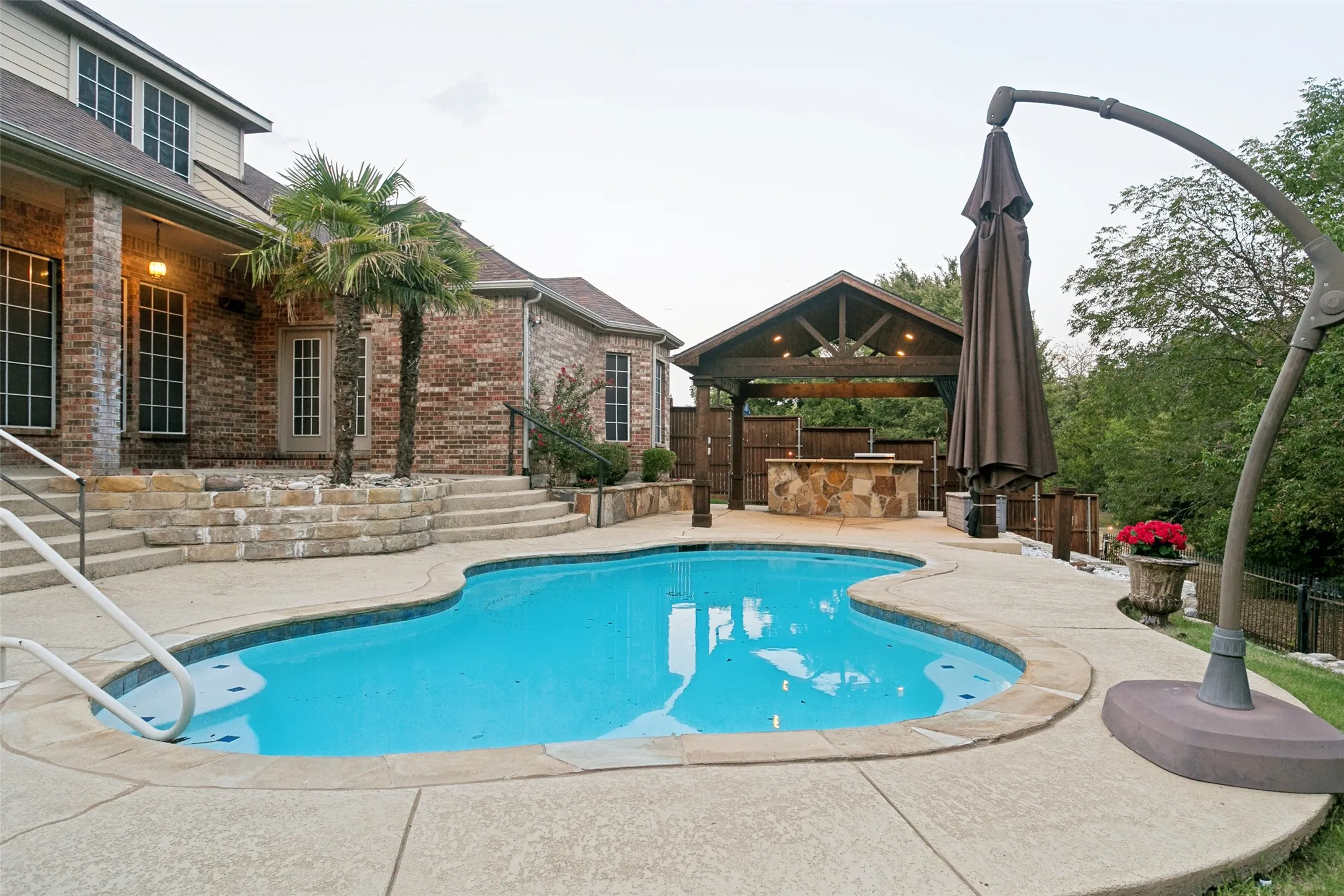 Curved swimming pool with surrounding concrete decking, a covered outdoor kitchen featuring stone veneer, and a brick exterior with multiple windows