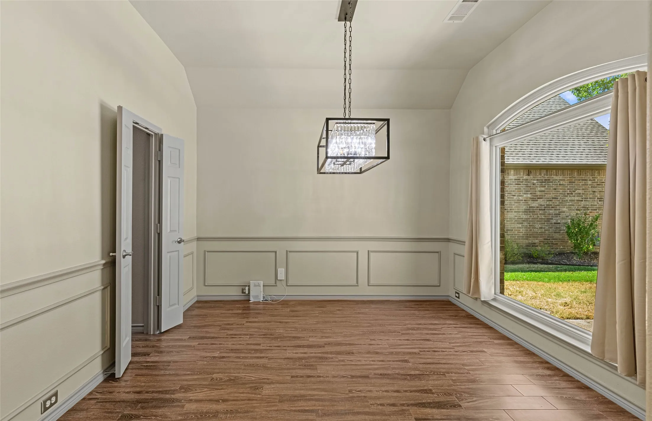 Wood-finish flooring, wainscoting, vaulted ceiling, arched window, and a chandelier with a crystal accent