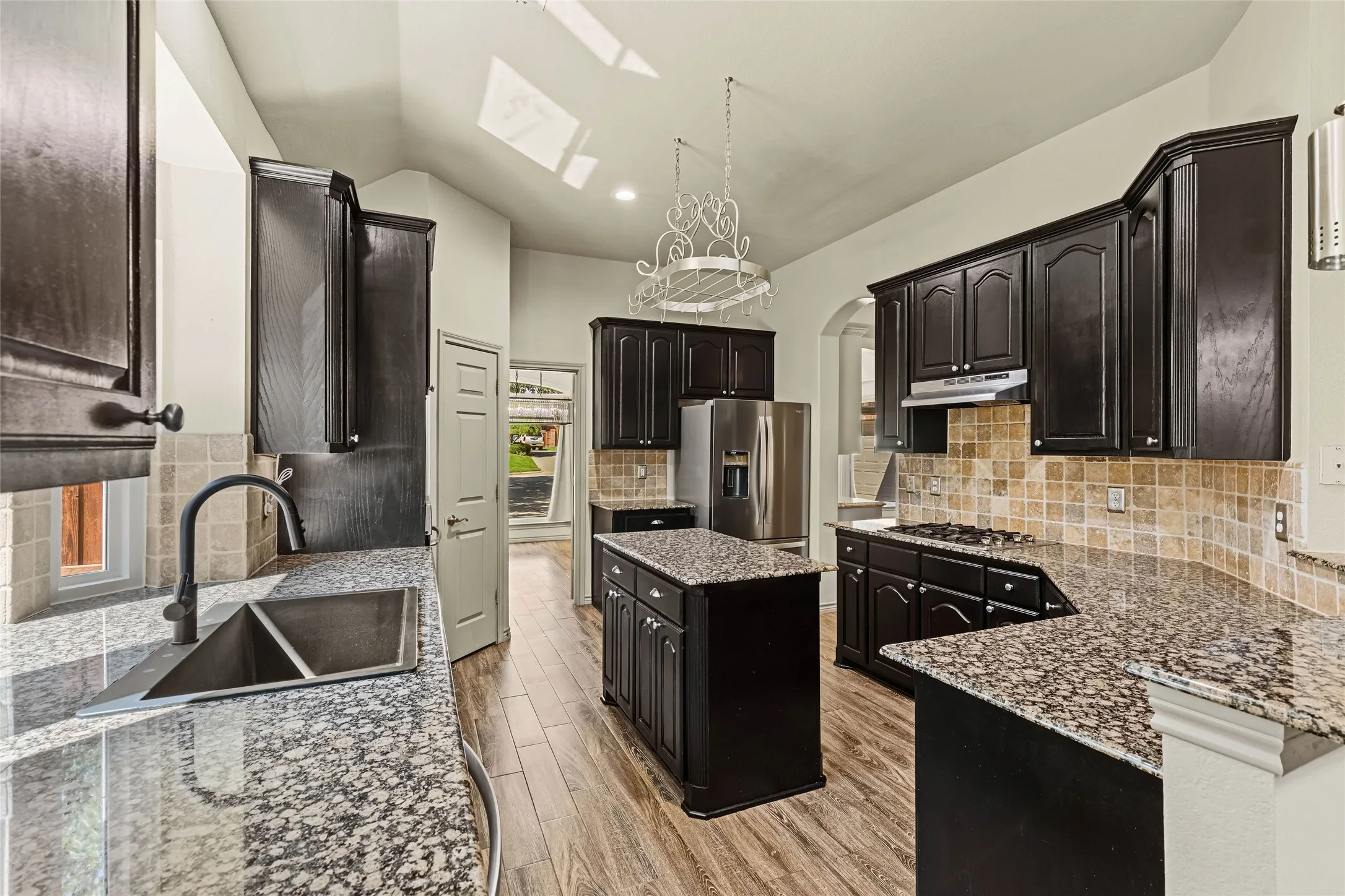 Kitchen featuring dark wood-finish cabinetry, granite countertops, stone tile backsplash, a central island, and wood-finish flooring