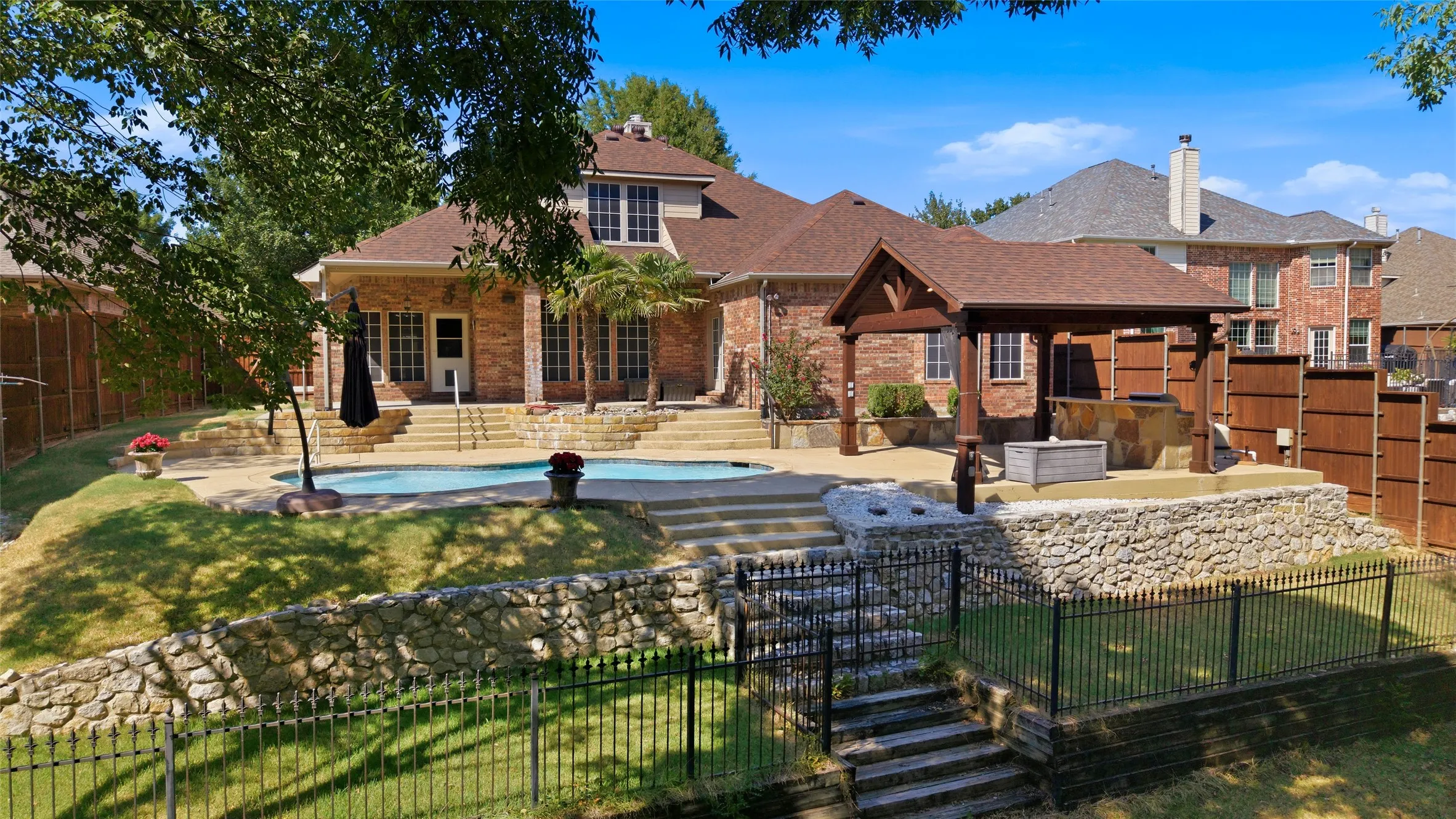 Brick exterior residence featuring a swimming pool, stone retaining walls, a covered outdoor structure with built-in features, and a fenced yard