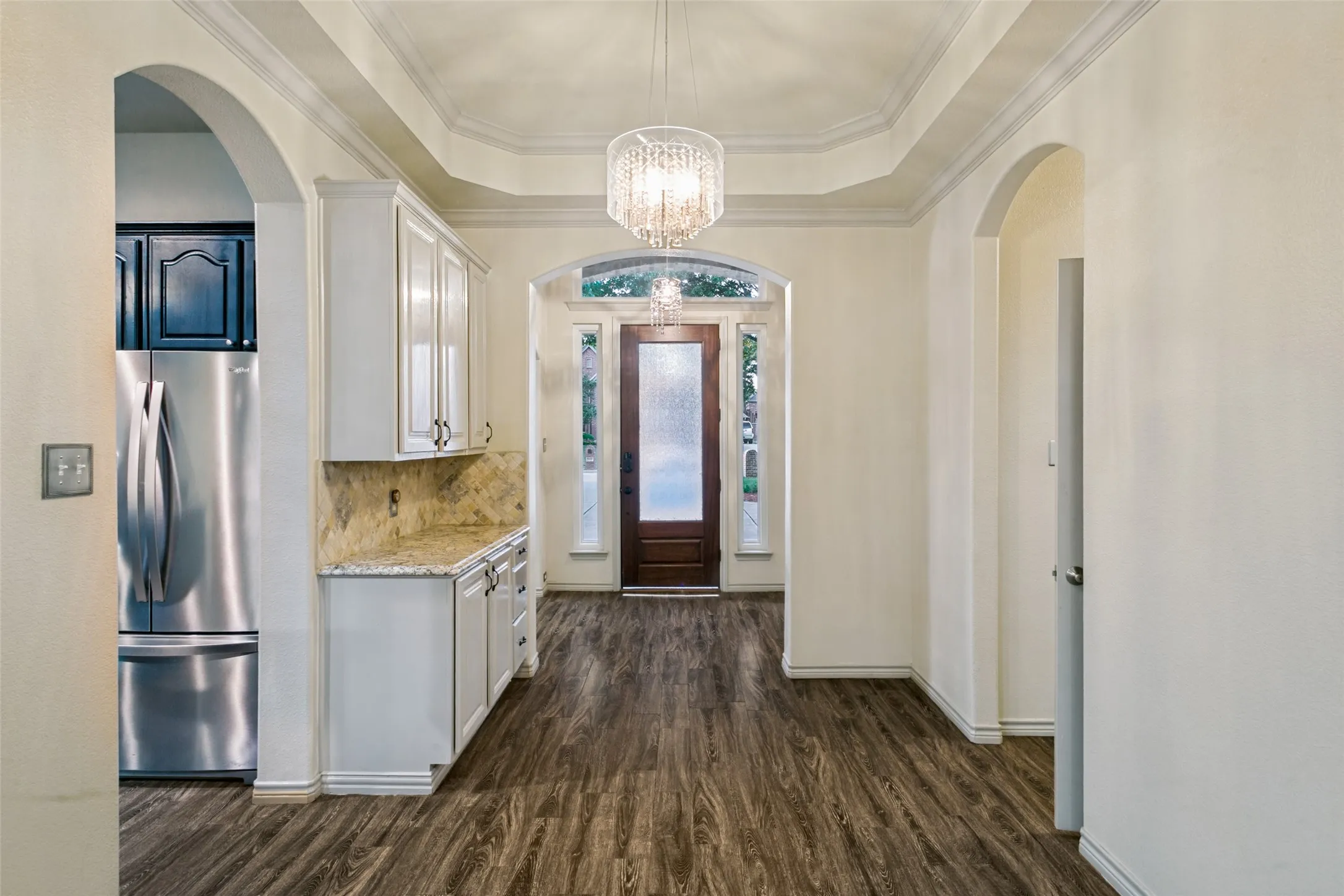Wood-finish flooring throughout, tray ceiling with a chandelier, archways, white cabinetry with a stone backsplash, and a brown paneled door with glass accents