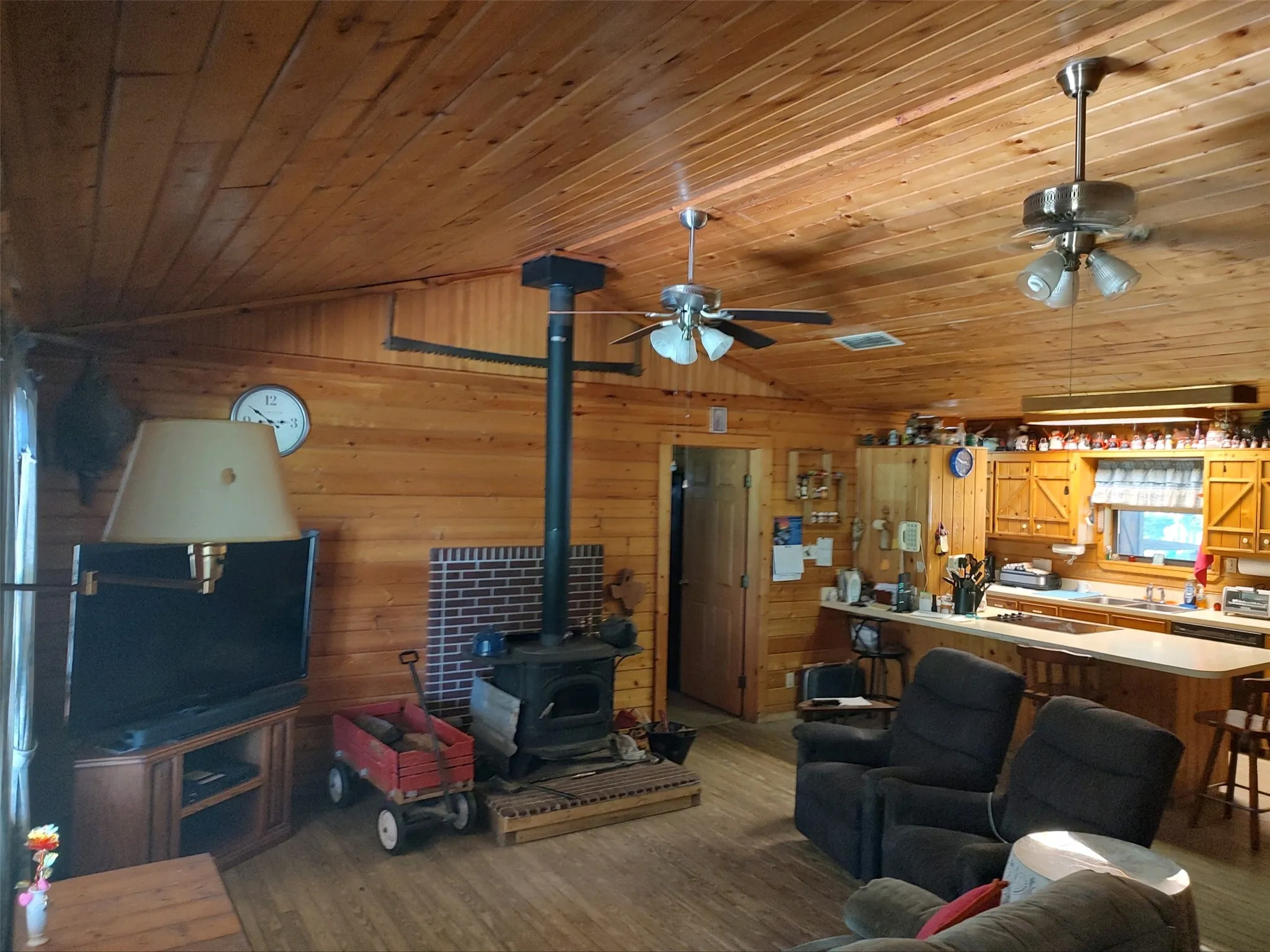 Living room, wood walls and ceilings, wood flooring, wood stove, open floor plan, bar with cooktop, view looking toward primary bedroom and kitchen.
