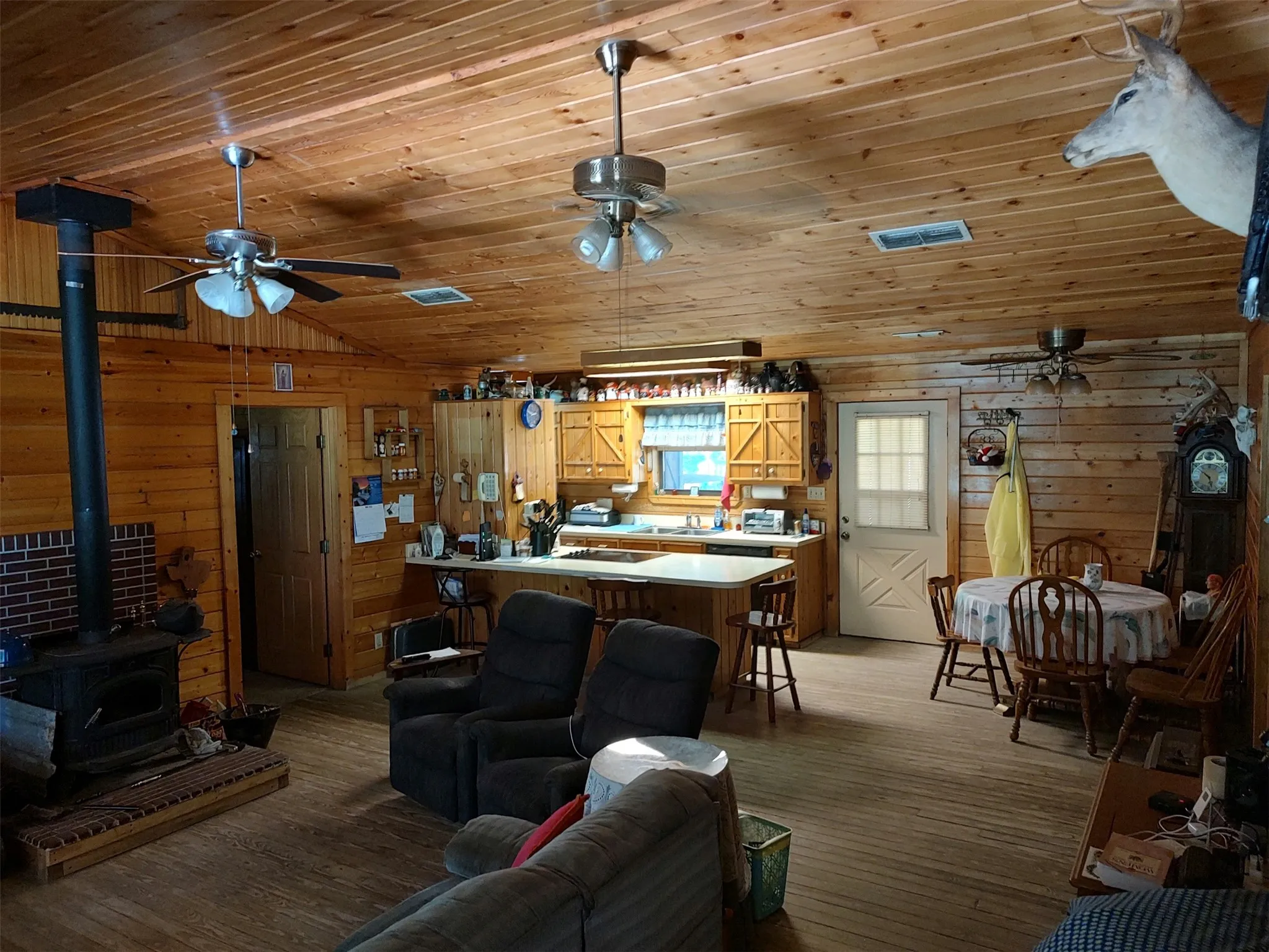 Living room, wood walls and ceilings, wood flooring, wood stove, open floor plan, bar with cooktop, view looking toward dining area and kitchen, back door by dining and kitchen.