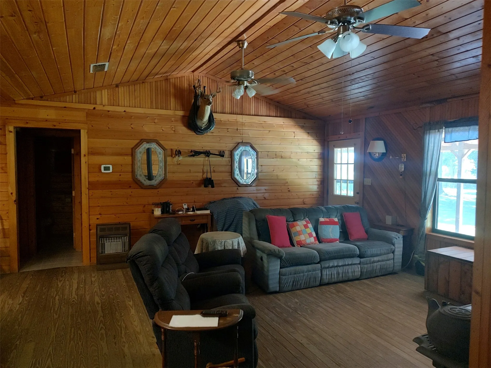 Living room, wood walls and ceilings, wood flooring, wood stove, open floor plan, view looking from primary bedroom, front door in corner.