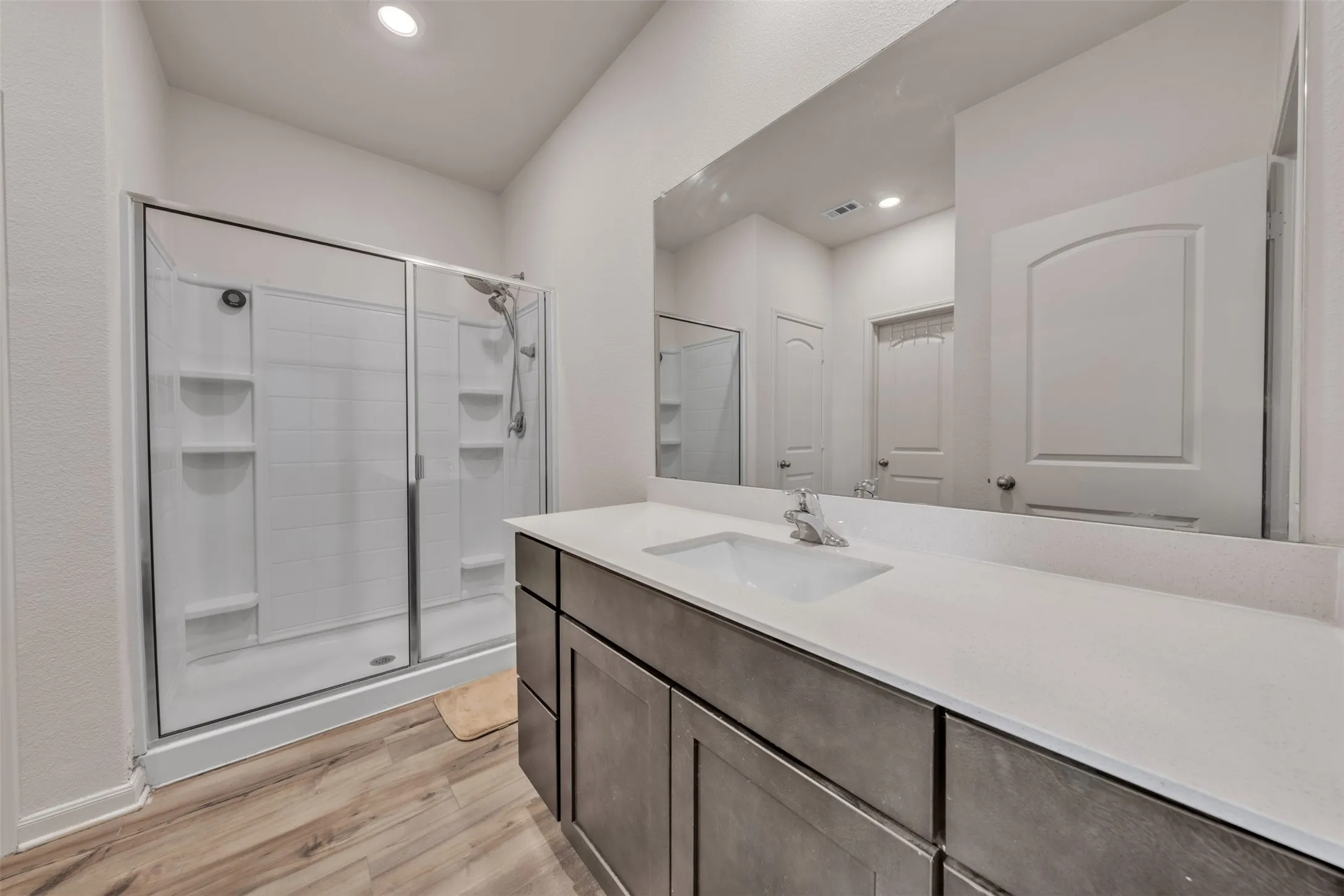 Wood-finish flooring, a spacious shower with clear glass enclosure, an extended vanity with dual sinks, and a large wall-mounted mirror