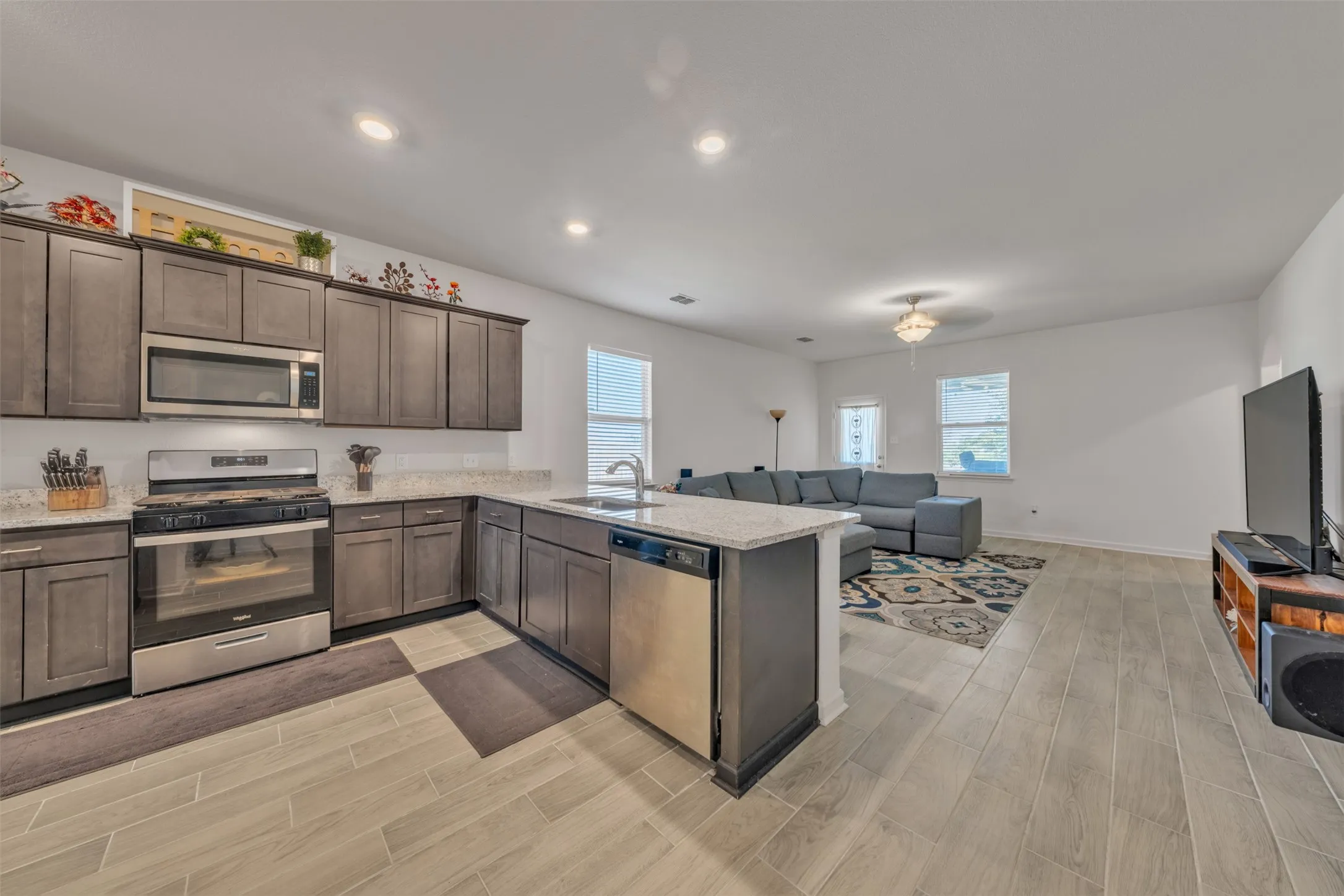 Open-concept living area with wood-finish flooring, recessed lighting, and a well-appointed kitchen featuring stainless steel appliances, grey cabinetry, and light-colored countertops