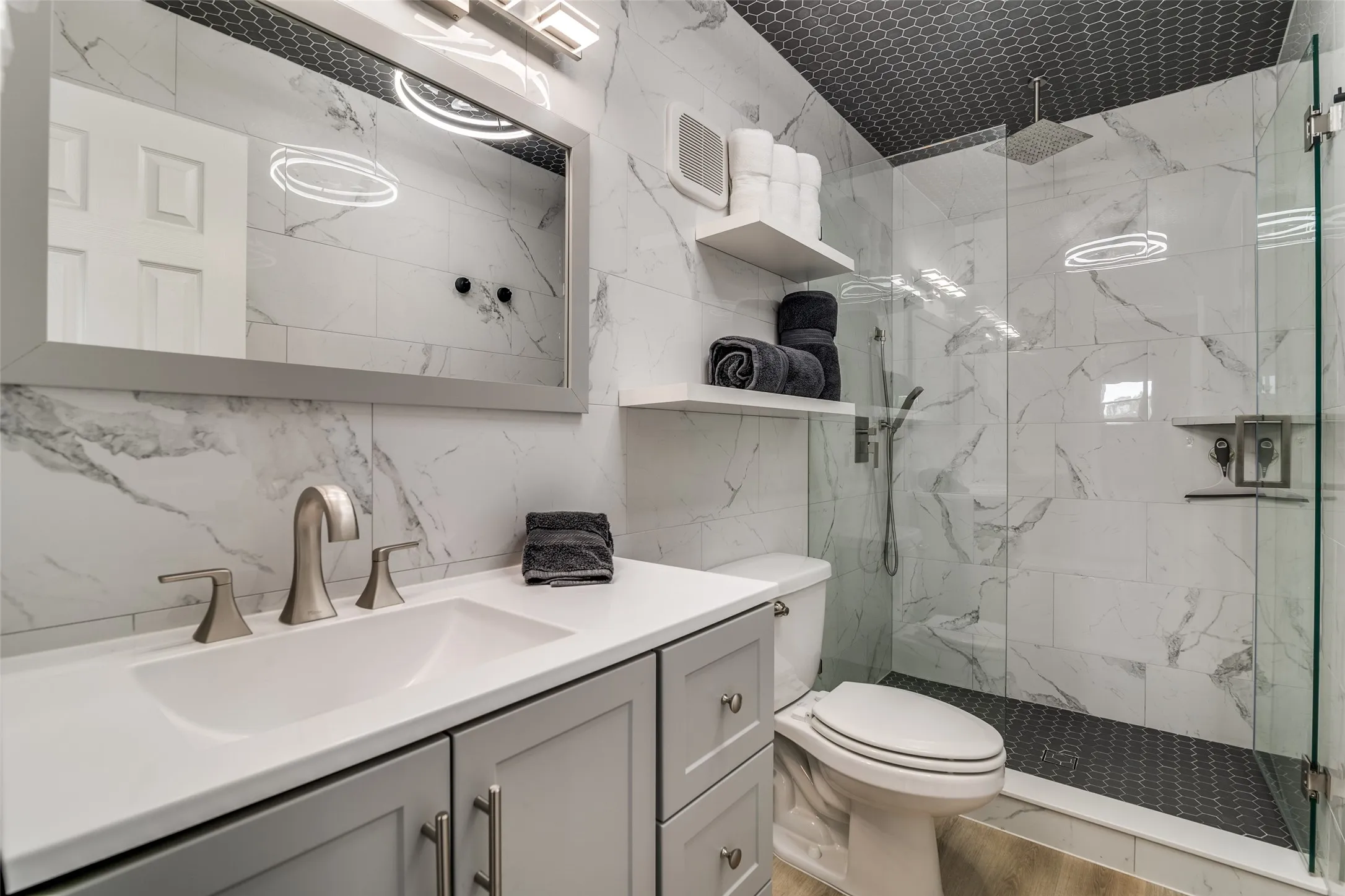 White vanity with an integrated sink and brushed nickel faucet, large framed mirror with overhead lighting, white marble-look tile walls, glass-enclosed shower with a rainfall showerhead, and a dark hexagonal tile shower floor
