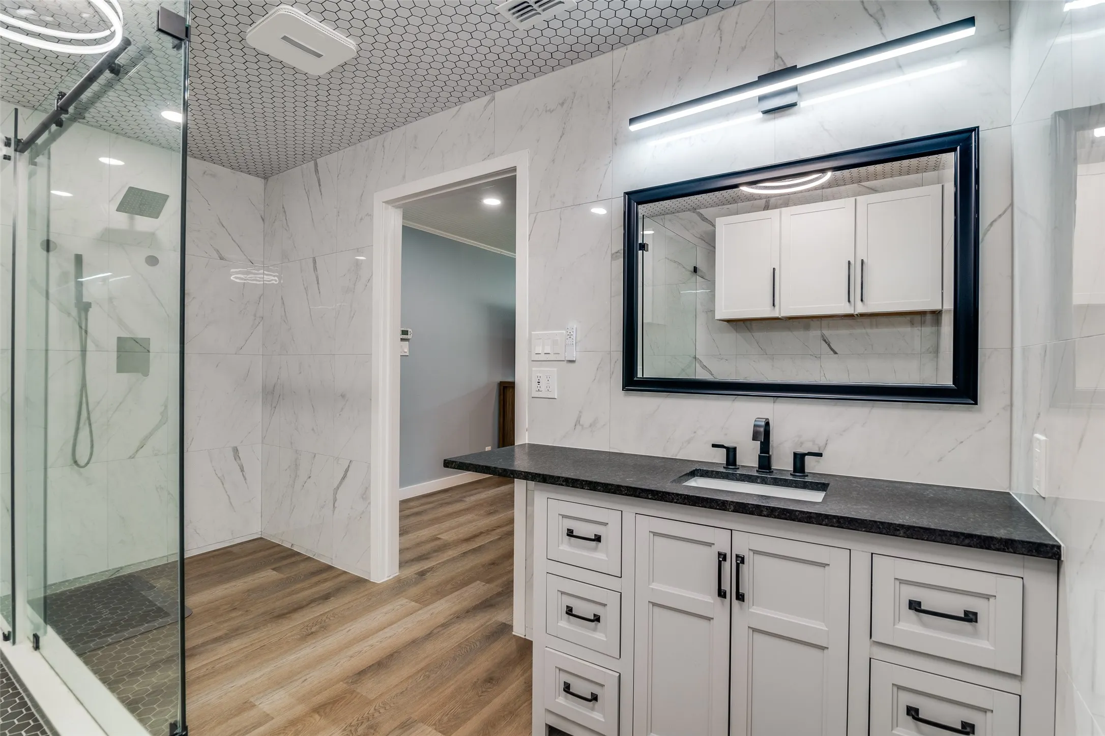 Glass-enclosed shower with wall-mounted fixtures, white hexagon tile ceiling, wood-finish flooring, white vanity with multiple drawers, and a dark countertop with an undermount sink