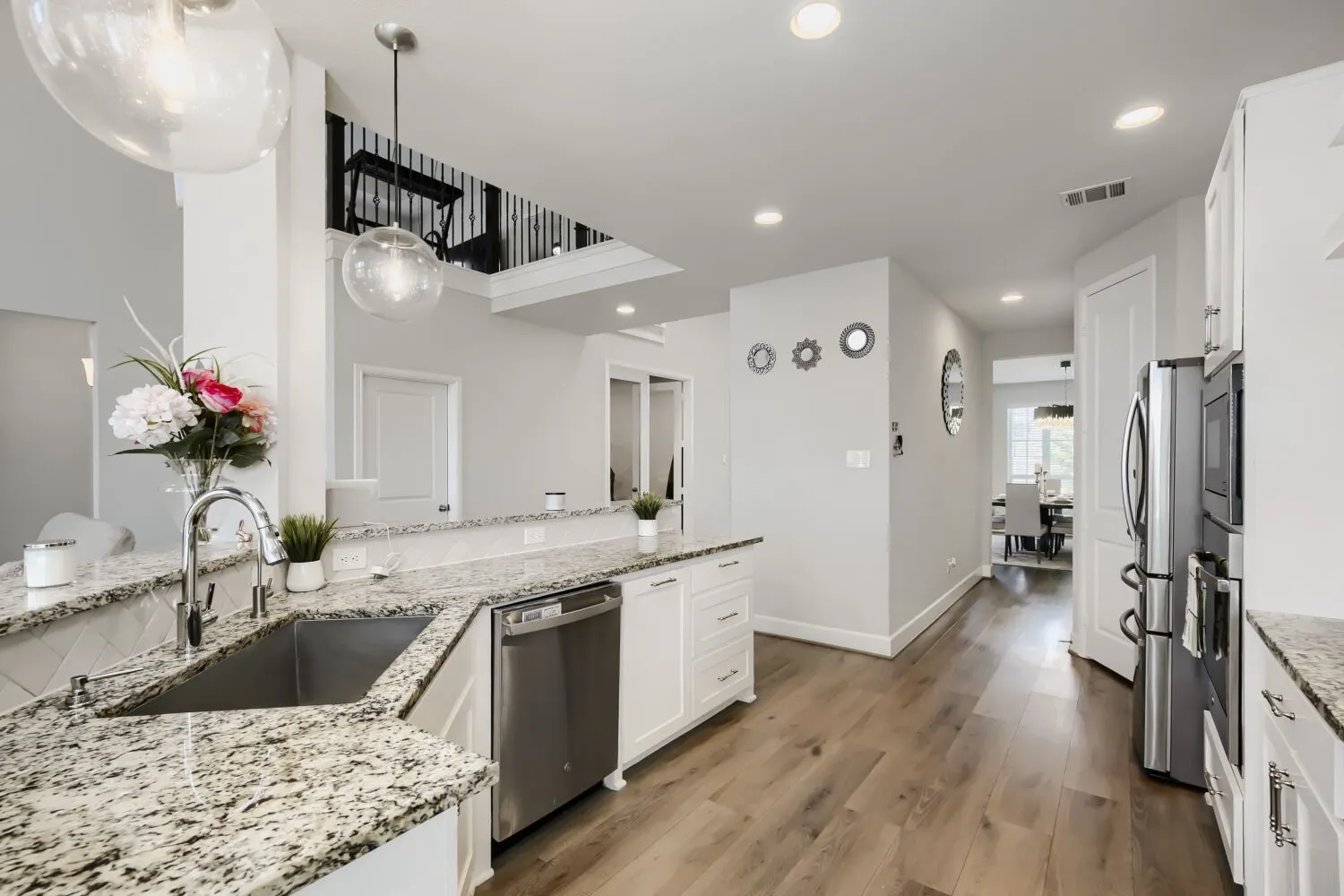 Modern kitchen featuring white cabinetry, granite-topped counters with waterfall edge, stainless steel appliances including a gas cooktop and under-mount sink, pendant lighting, and wide-plank wood-finish flooring throughout the open-concept space
