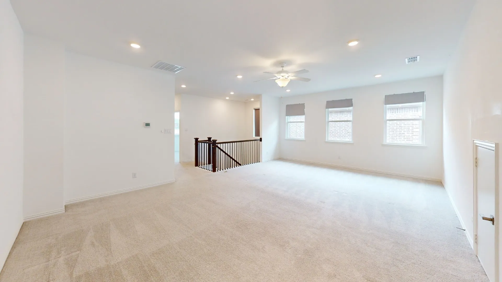 Expansive carpeted area featuring recessed lighting, a ceiling fan, a dark wood and wrought iron staircase railing, and three windows with shades
