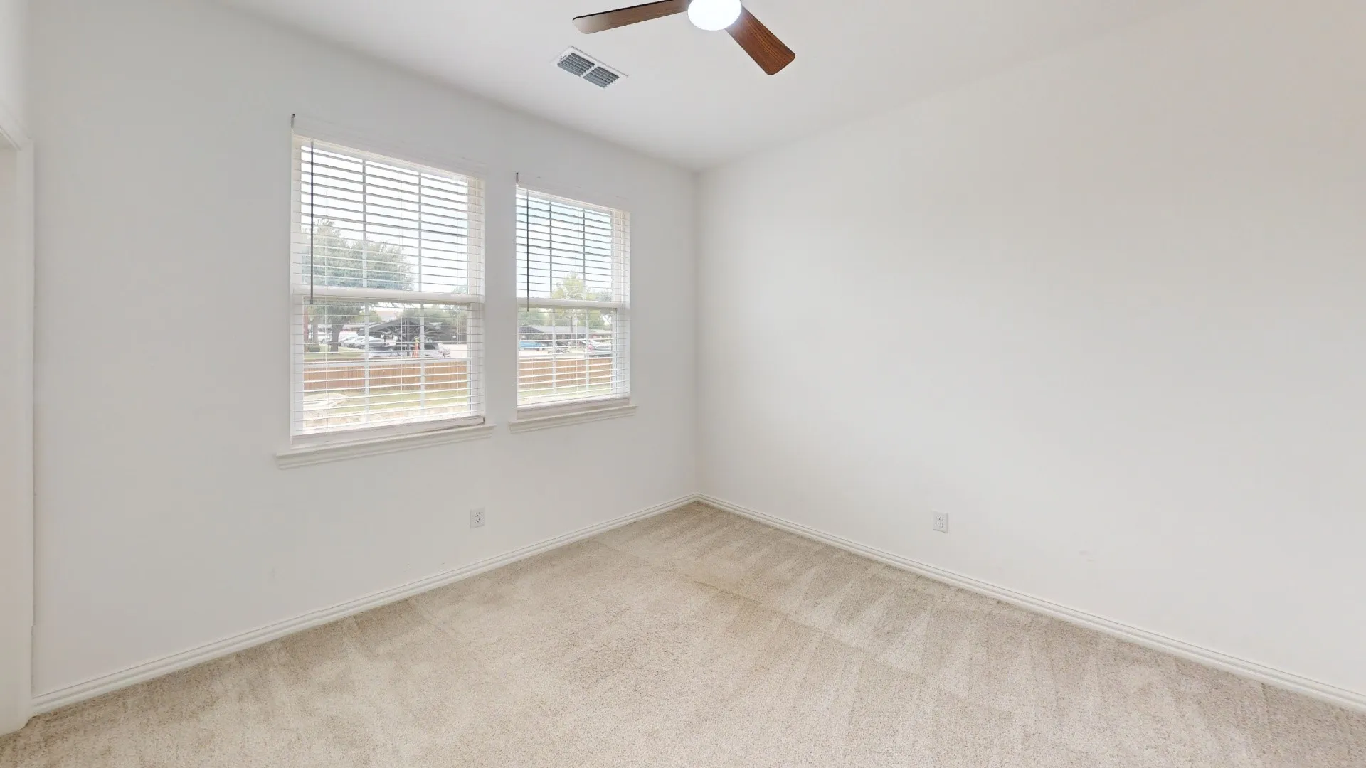 White walls, light-toned carpet, two windows with blinds, and a ceiling fan