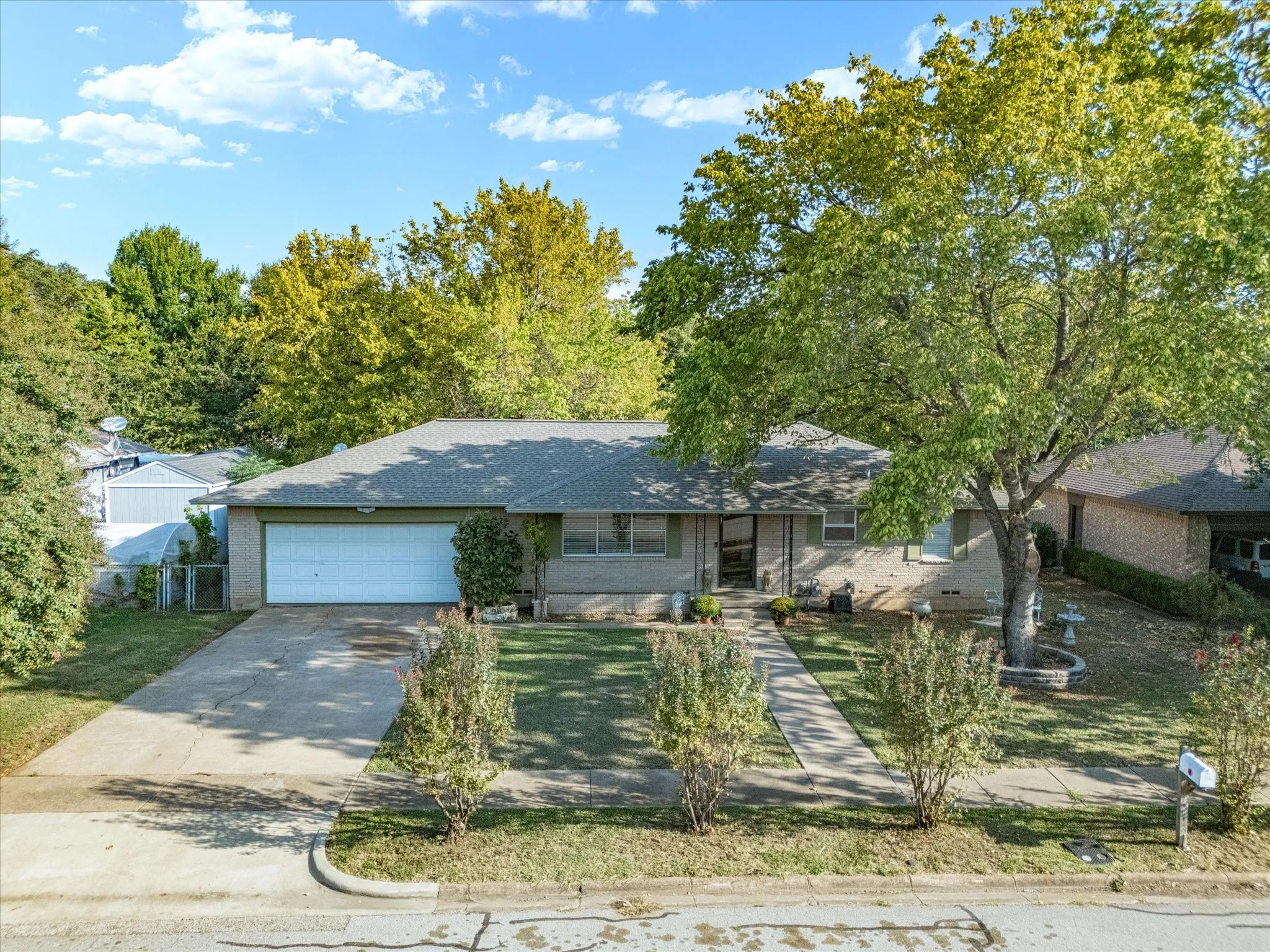 Single-story residence featuring a brick exterior, attached garage, concrete driveway, and mature landscaping with established trees