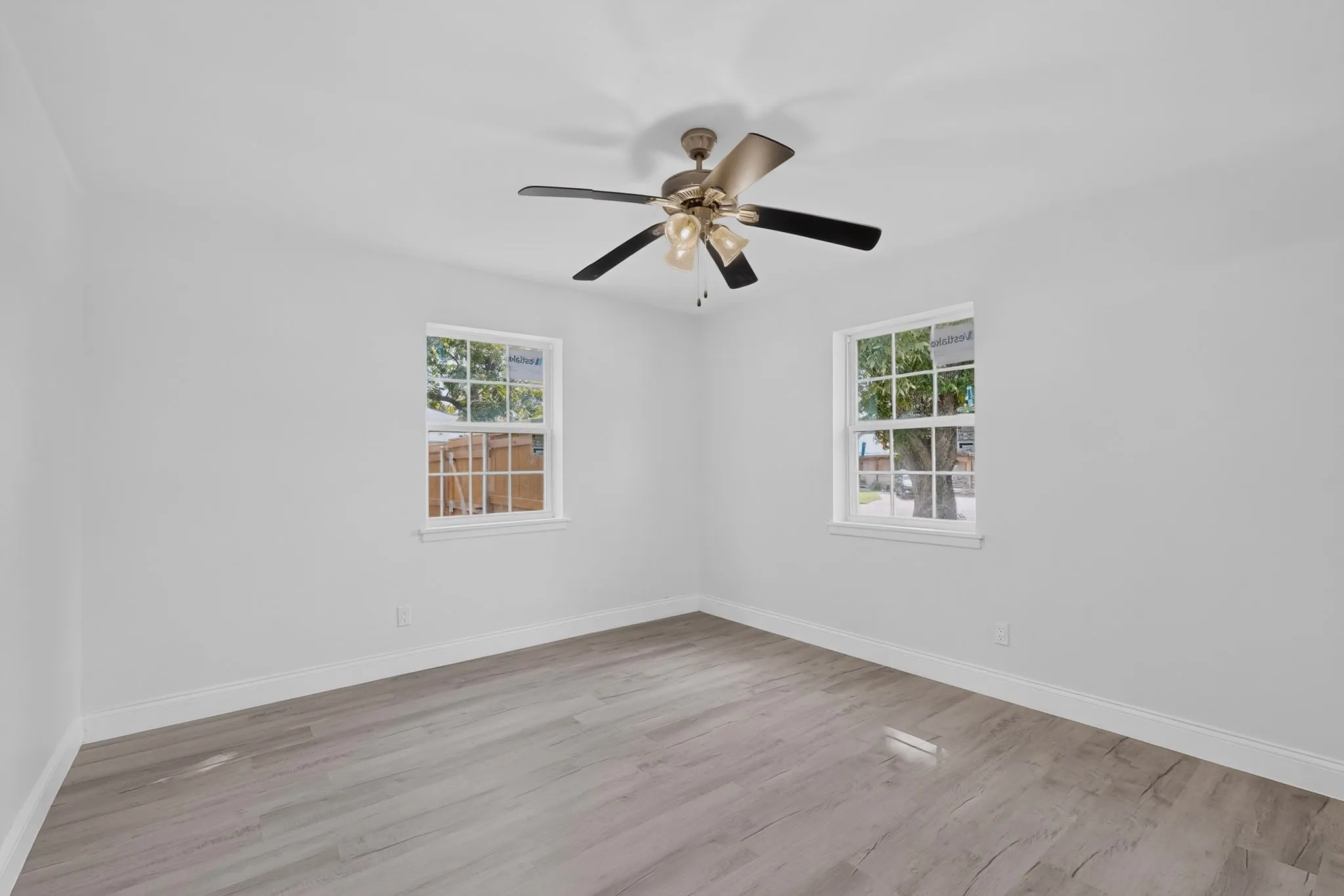 Spacious room featuring wood-finish flooring, white walls, two large windows, and a ceiling fan with integrated lighting