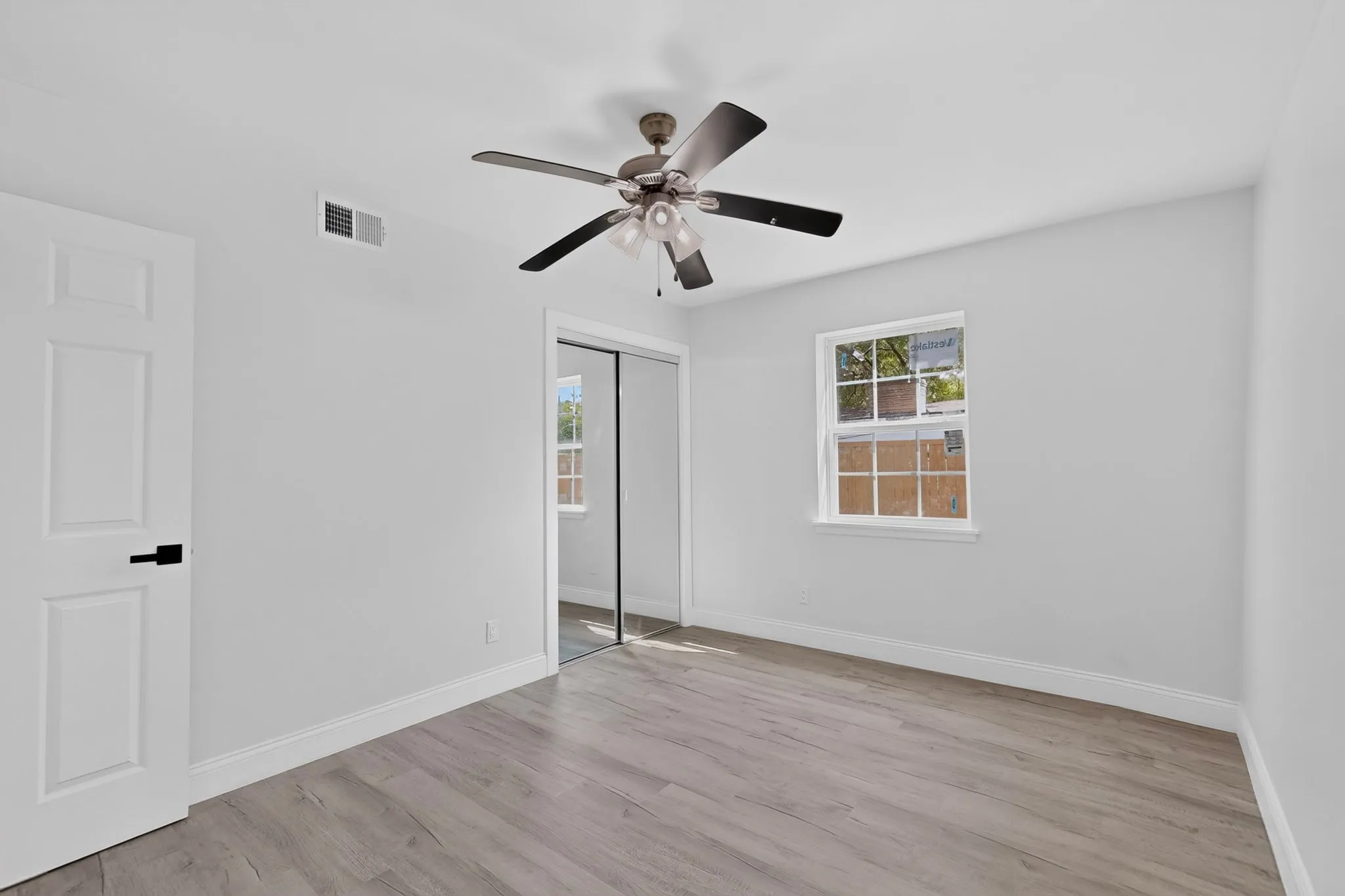 White wall paint, wood-finish flooring, ceiling fan with light, white trim, and a window with white casing, mirrored closet doors
