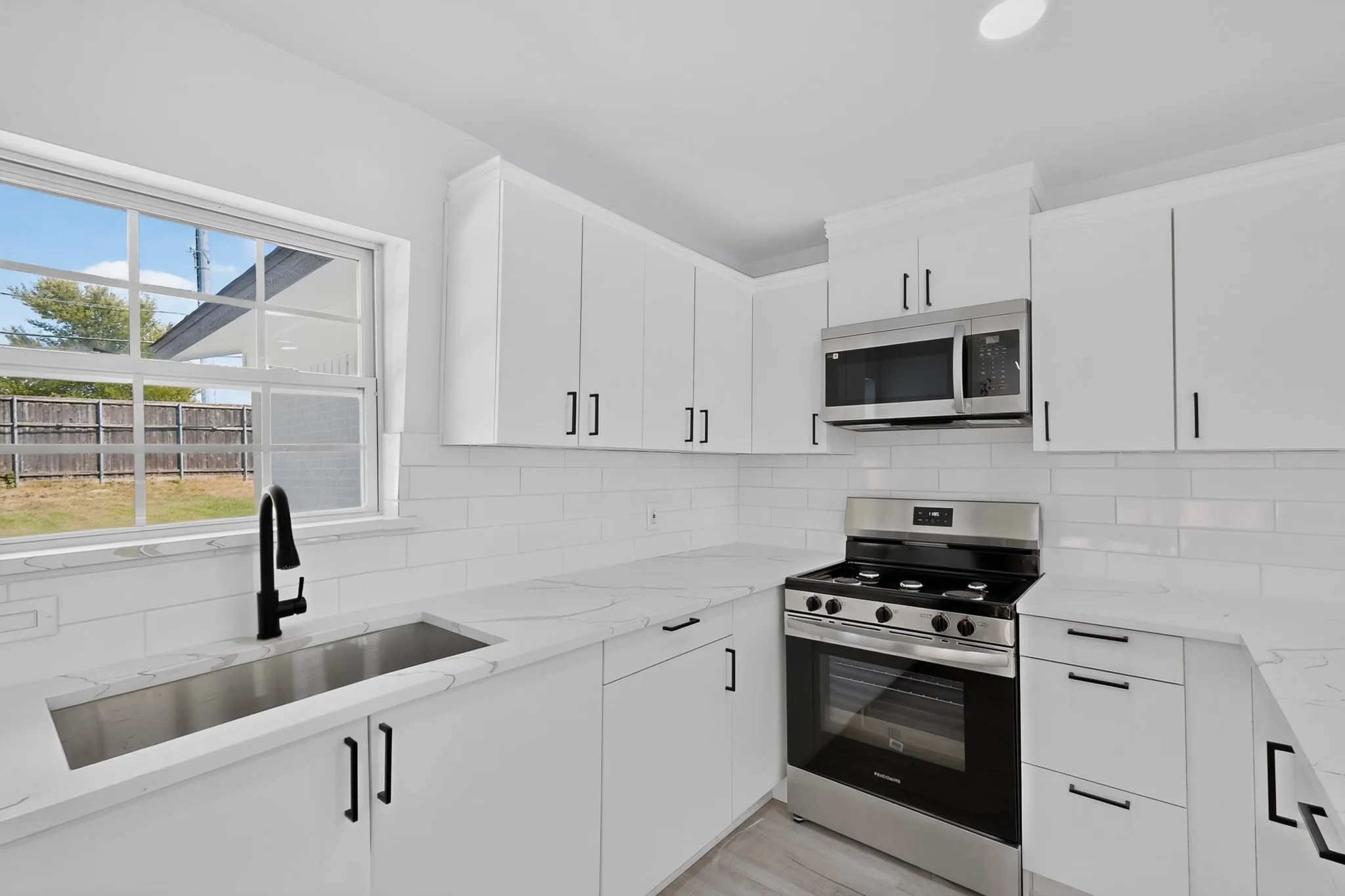 Updated kitchen featuring white shaker cabinetry, stainless steel appliances, a stainless steel undermount sink with a matte black faucet, white subway tile backsplash, and light-toned countertops