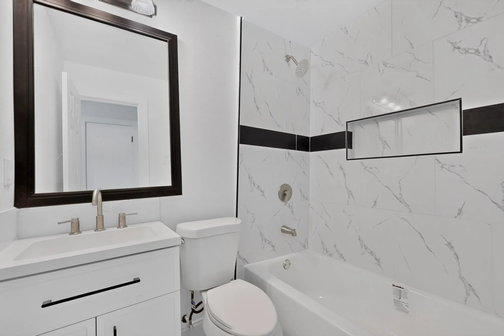 White vanity with brushed nickel faucet, large framed mirror, patterned tile shower surround, built-in shower niche, and white bathtub