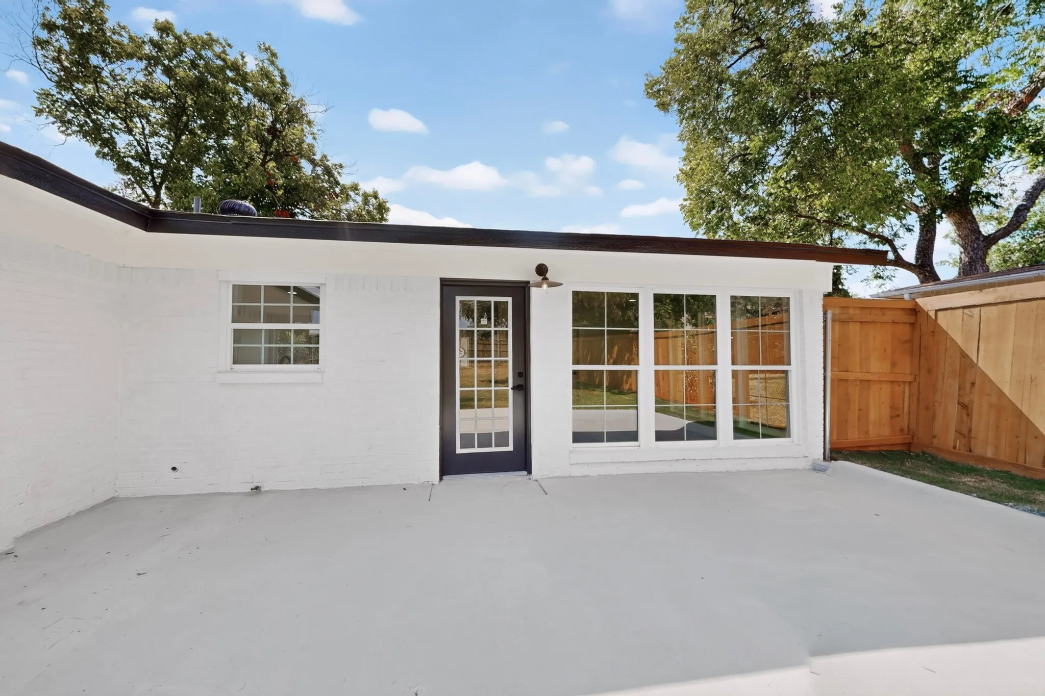 Exterior brick painted white, large concrete patio, dark frame glass-panel door, multiple large windows, and a wood fence