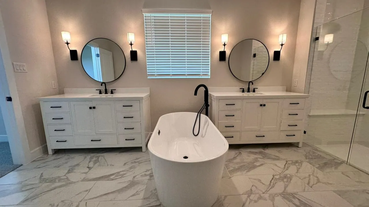 Freestanding soaking tub, dual vanity with white cabinetry, black matte fixtures, round mirrors, and a glass-enclosed shower with a bench