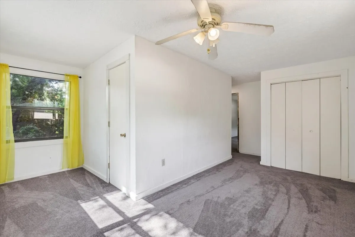 Carpeted flooring, white walls, ceiling fan with light fixture, window with exterior foliage, bi-fold closet doors