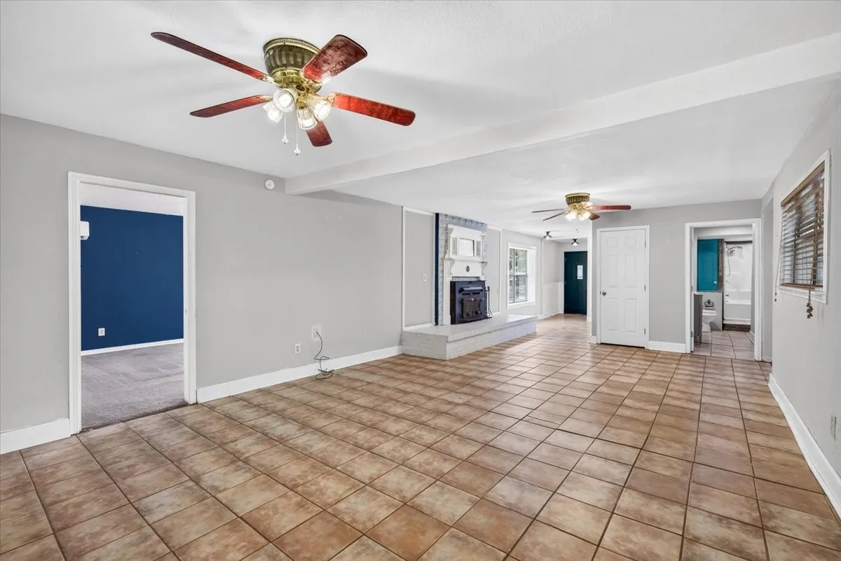 Expansive tiled flooring throughout, complemented by a built-in fireplace with a hearth, multiple ceiling fans, and a window with blinds