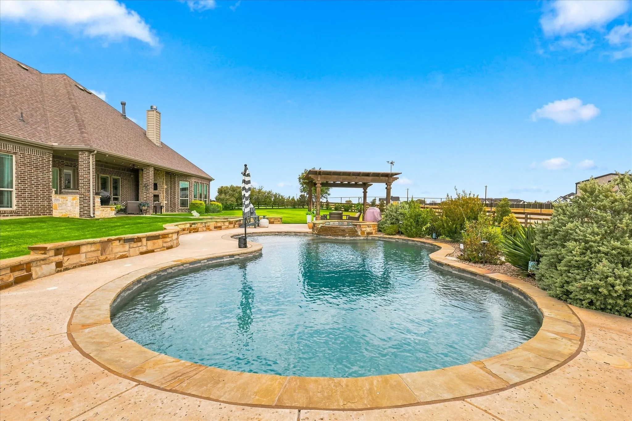 Curved swimming pool with stone coping, an adjacent stone paver patio, a wooden pergola, and a brick-clad residence