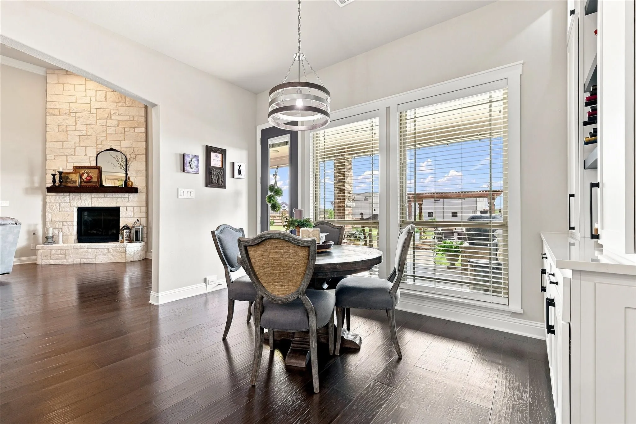Dark wood-finish flooring, stone fireplace with a wood mantle, white built-in cabinetry, large windows with blinds, and a contemporary chandelier