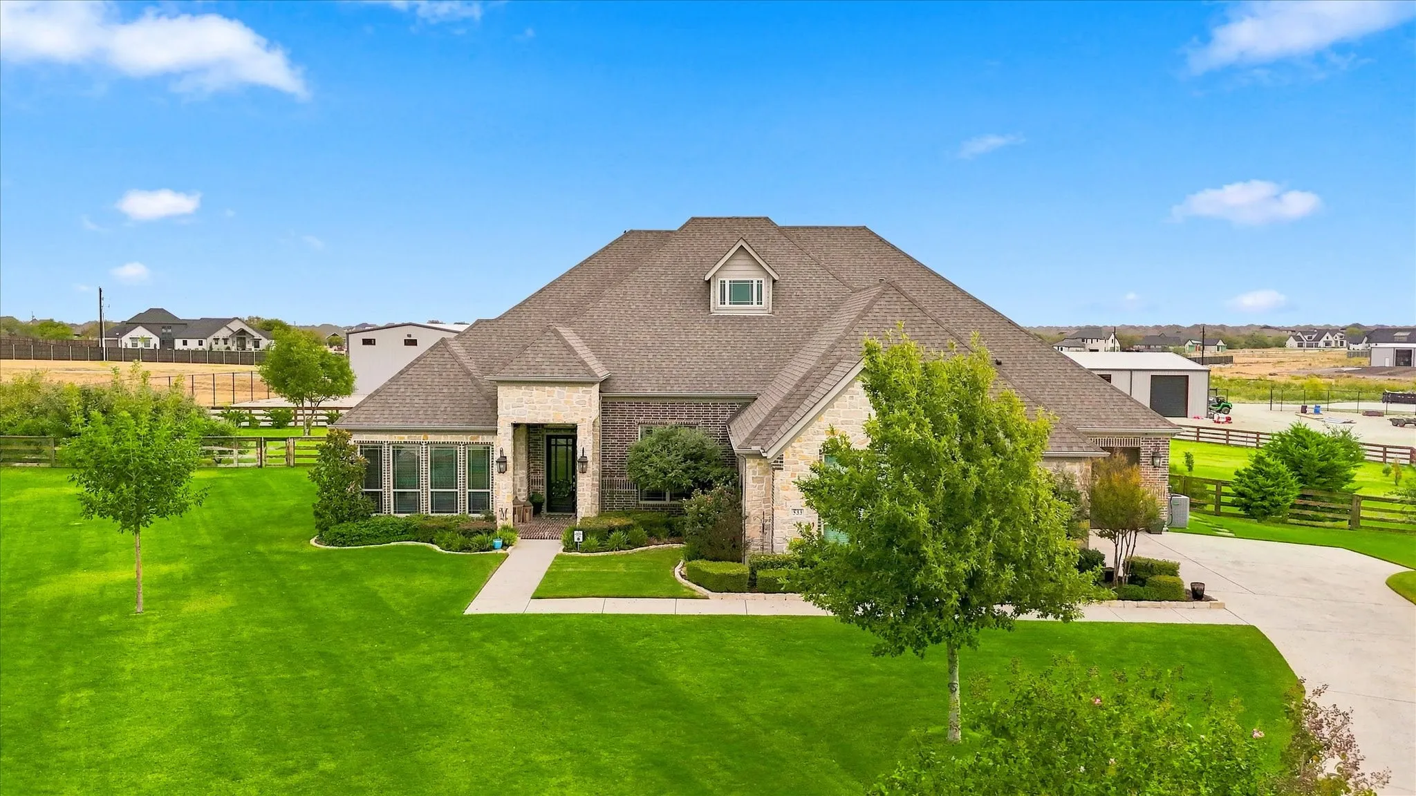 Brick and stone exterior with dormer window, covered entry, manicured landscaping, and expansive green lawn