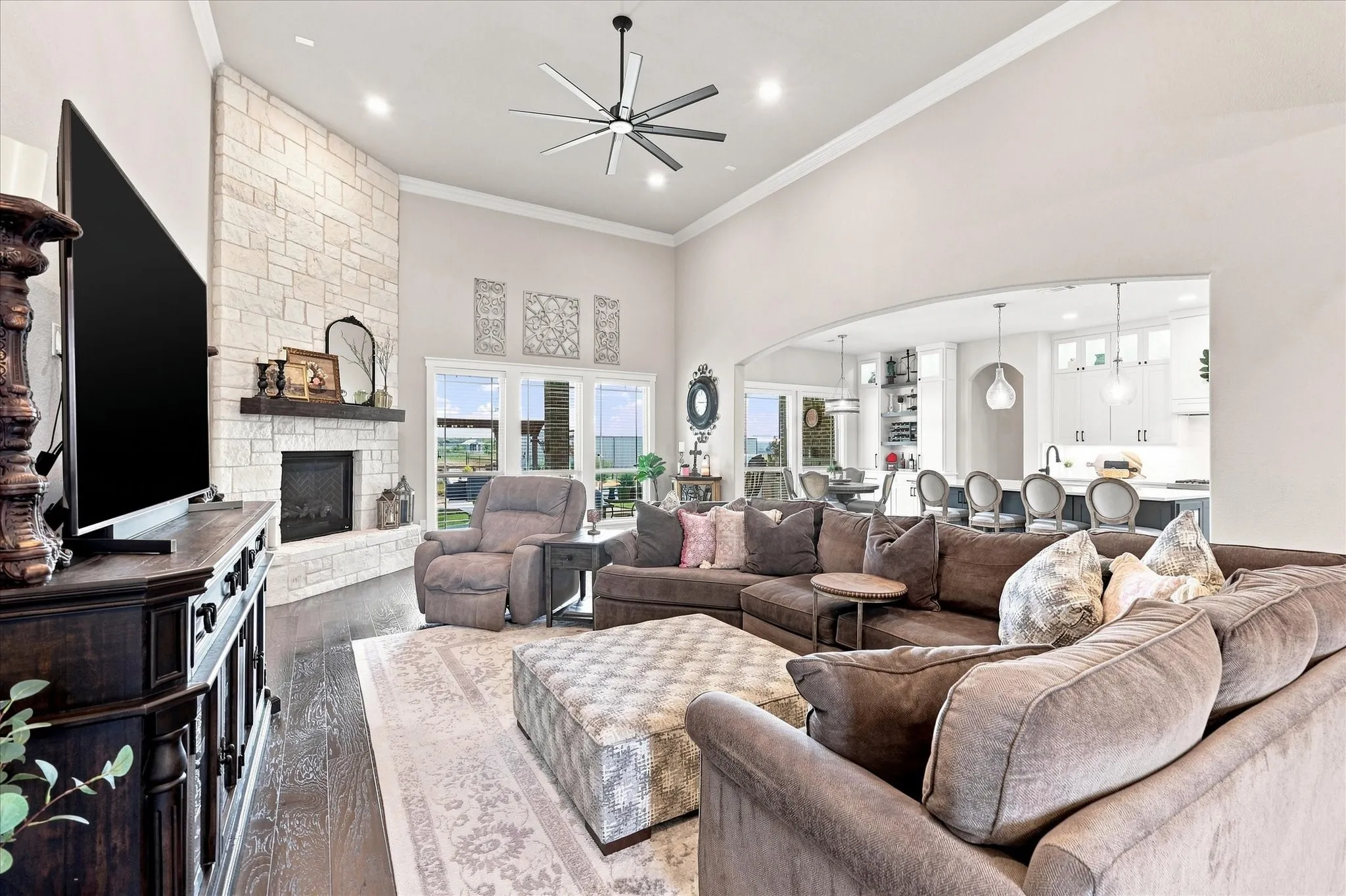 Vaulted ceiling with a contemporary ceiling fan, stone-clad fireplace with a dark wood mantel, dark wood-finish flooring, multiple windows, and an arched entryway to the kitchen