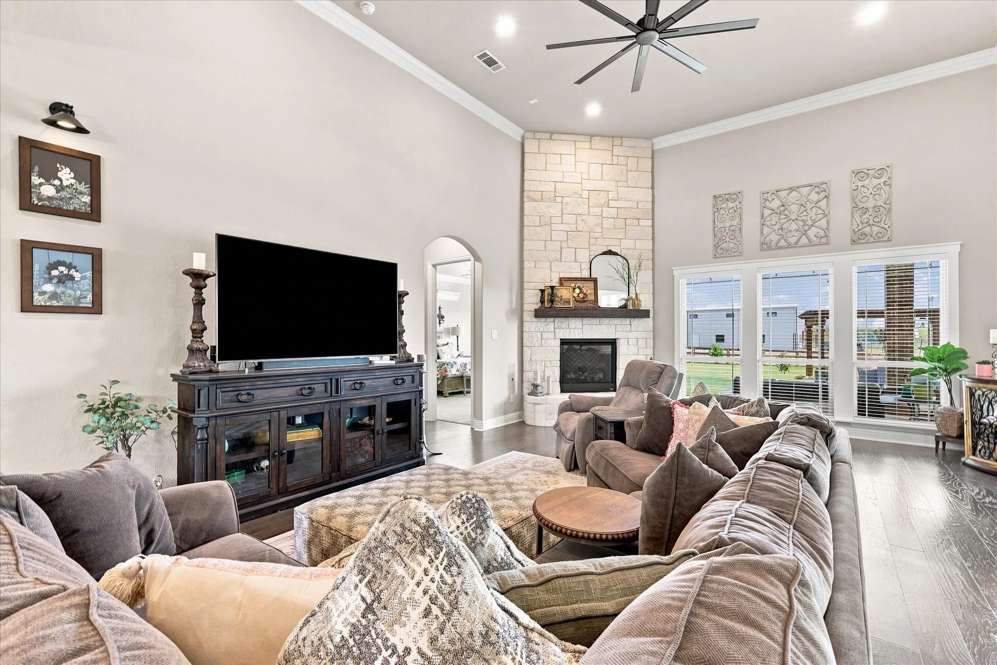 Vaulted ceiling with crown molding, stone fireplace with wood mantle, dark wood-finish flooring, recessed lighting, and large windows