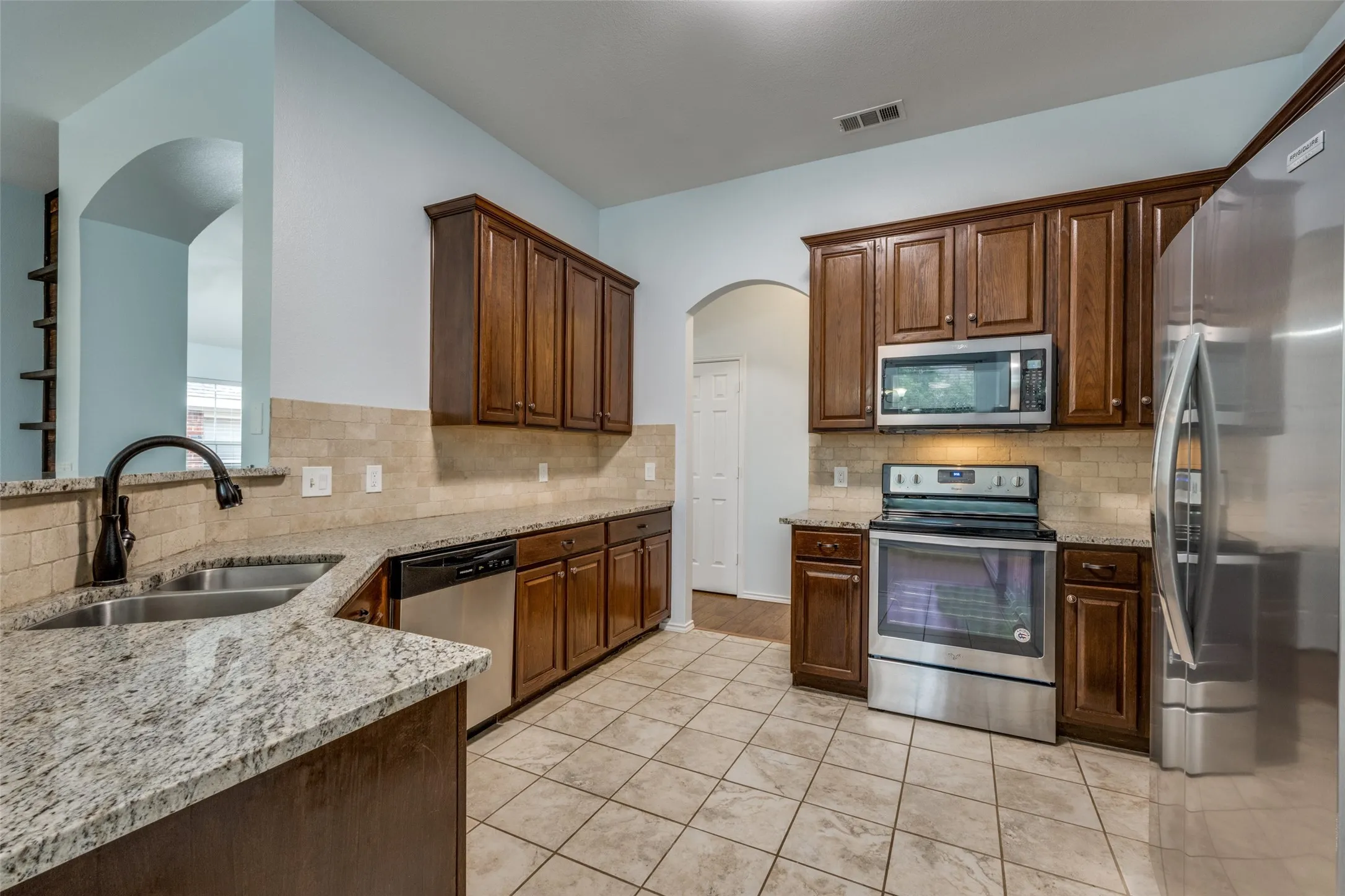 Kitchen featuring wood cabinetry, stainless steel appliances, granite countertops, tile backsplash, and tile flooring