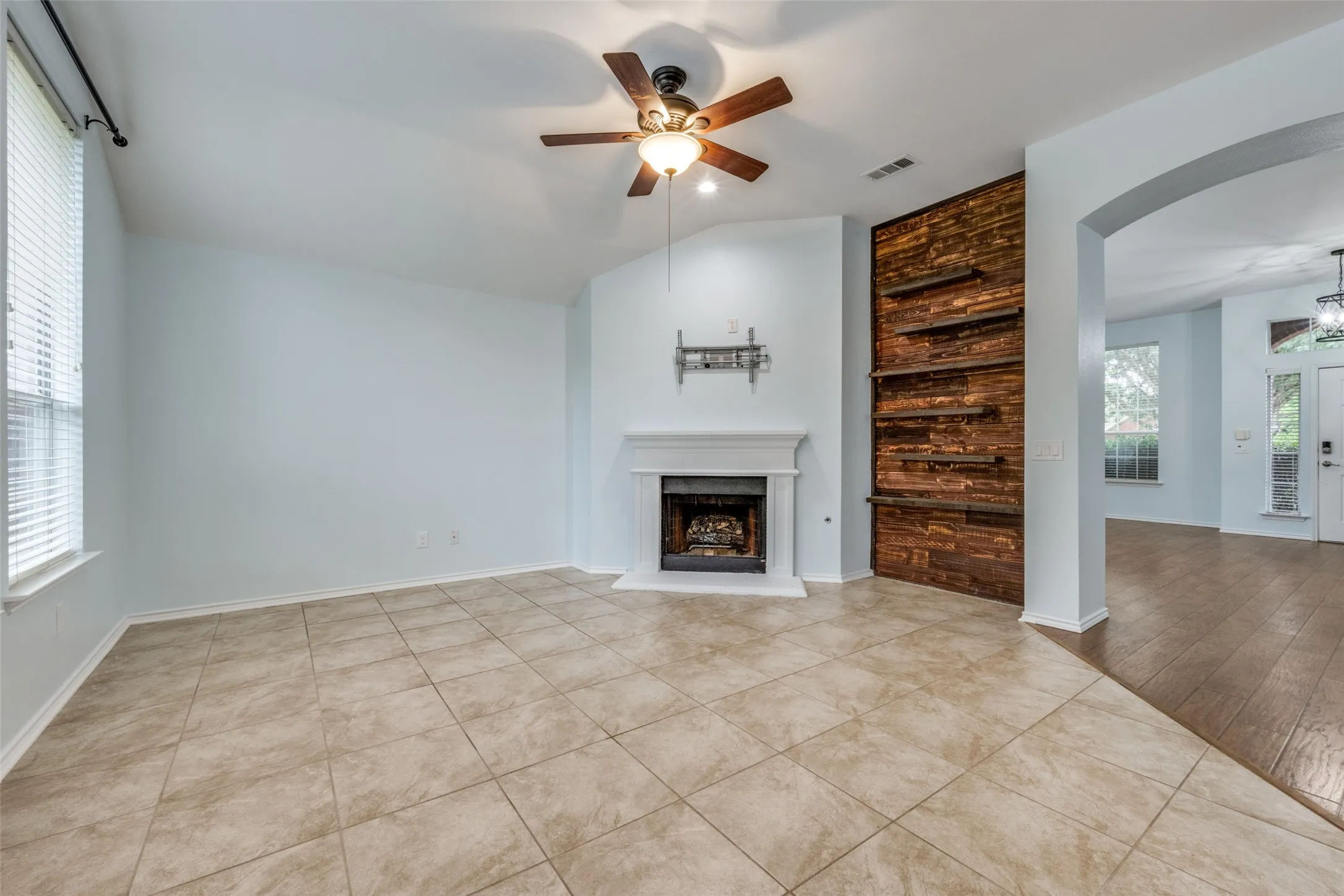 Tiled flooring, vaulted ceiling, fireplace with white mantel, wood-paneled accent wall with built-in shelving, and a ceiling fan with integrated lighting