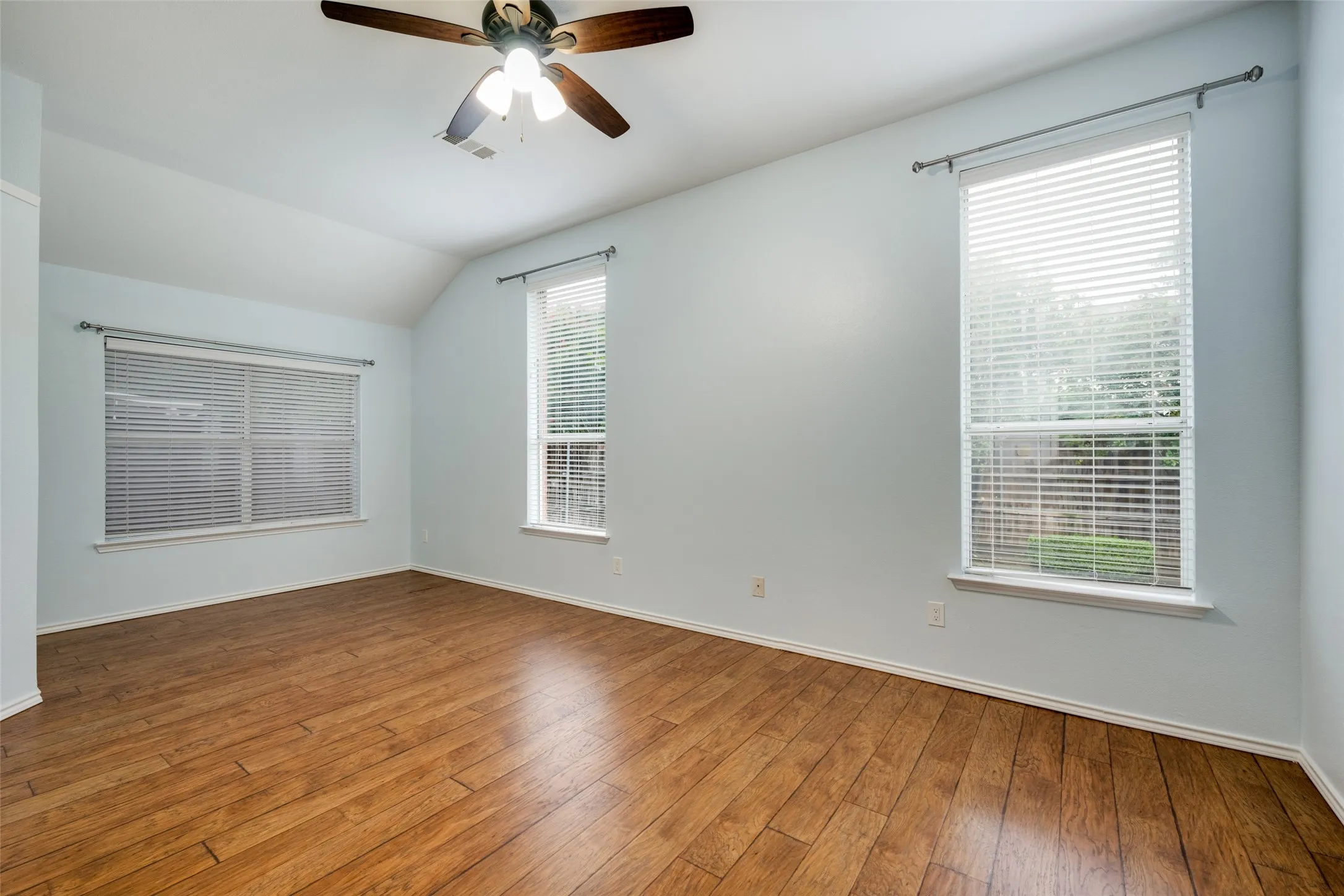 Vaulted ceiling with a ceiling fan, two large windows, wood-finish flooring, and light-colored walls