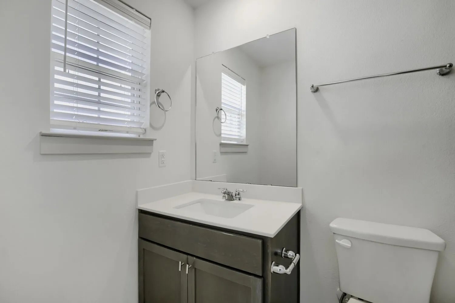 Vanity with a white countertop and integrated sink, a rectangular mirror, and a window with blinds