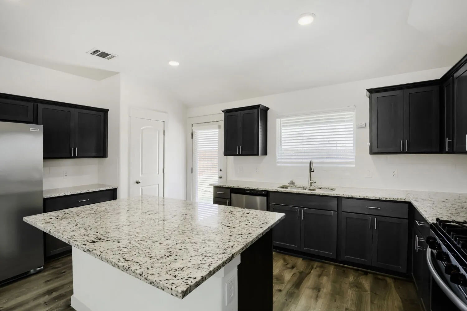 Kitchen featuring dark shaker-style cabinetry, light speckled countertops, stainless steel appliances, a center island, and wood-finish flooring
