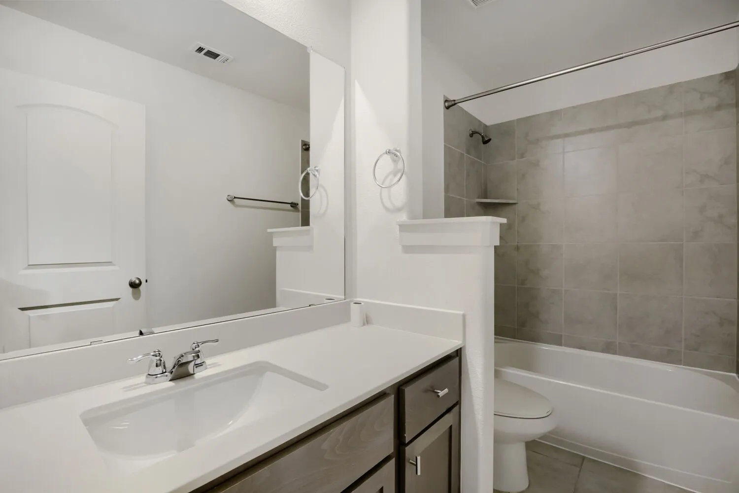 Vanity with integrated sink and chrome faucet, large frameless mirror, wood-finish cabinetry, tiled shower surround, and built-in shower shelf