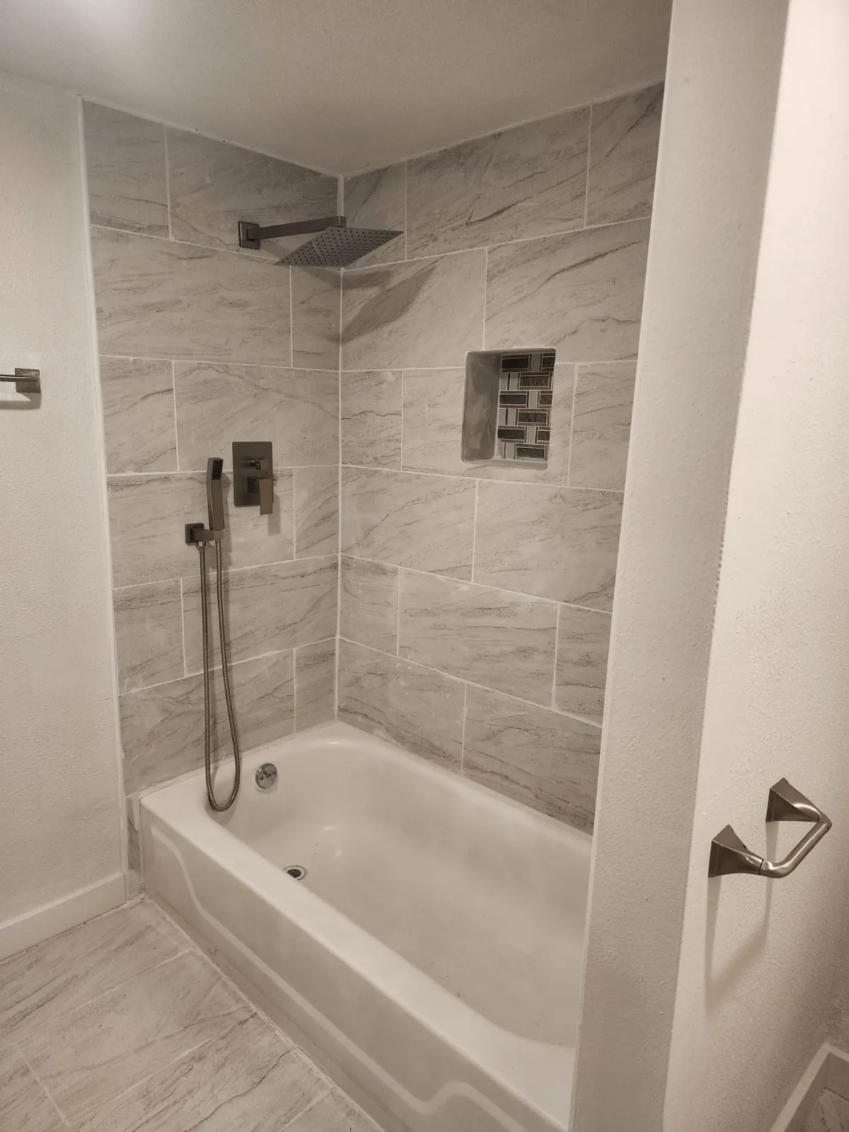 Bathtub with light-toned wall tile, a built-in shower niche with mosaic tile, a ceiling-mounted rainfall showerhead, and a handheld shower attachment