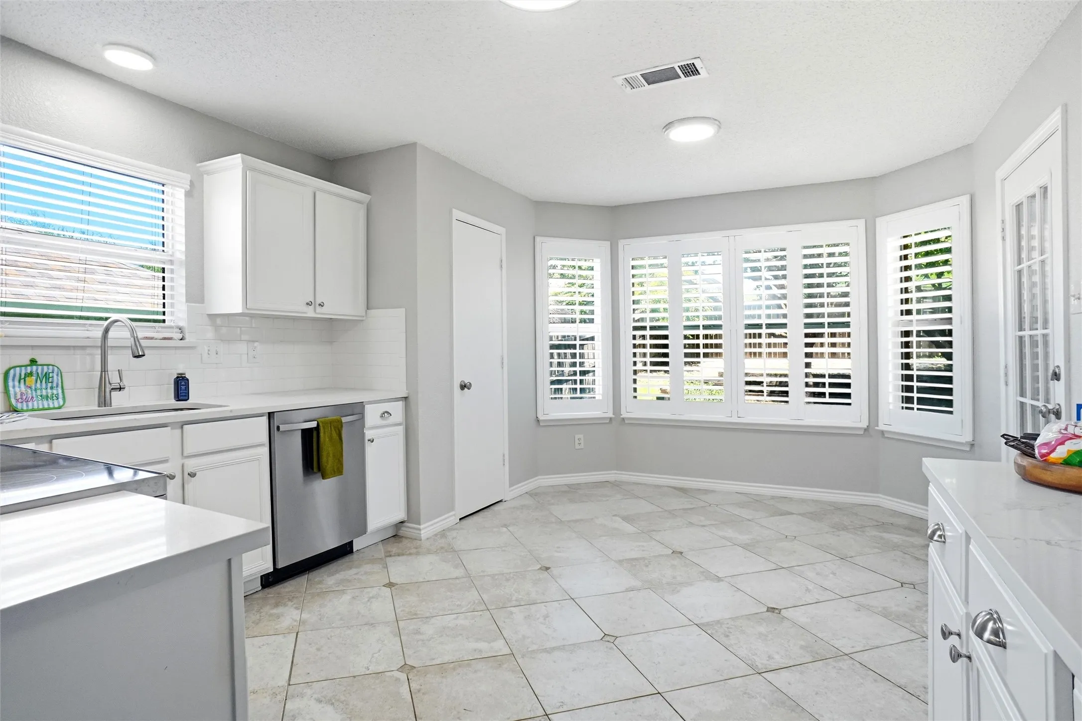 Kitchen featuring white cabinetry, light-toned countertops, a stainless steel dishwasher, tile flooring, and multiple windows with plantation shutters