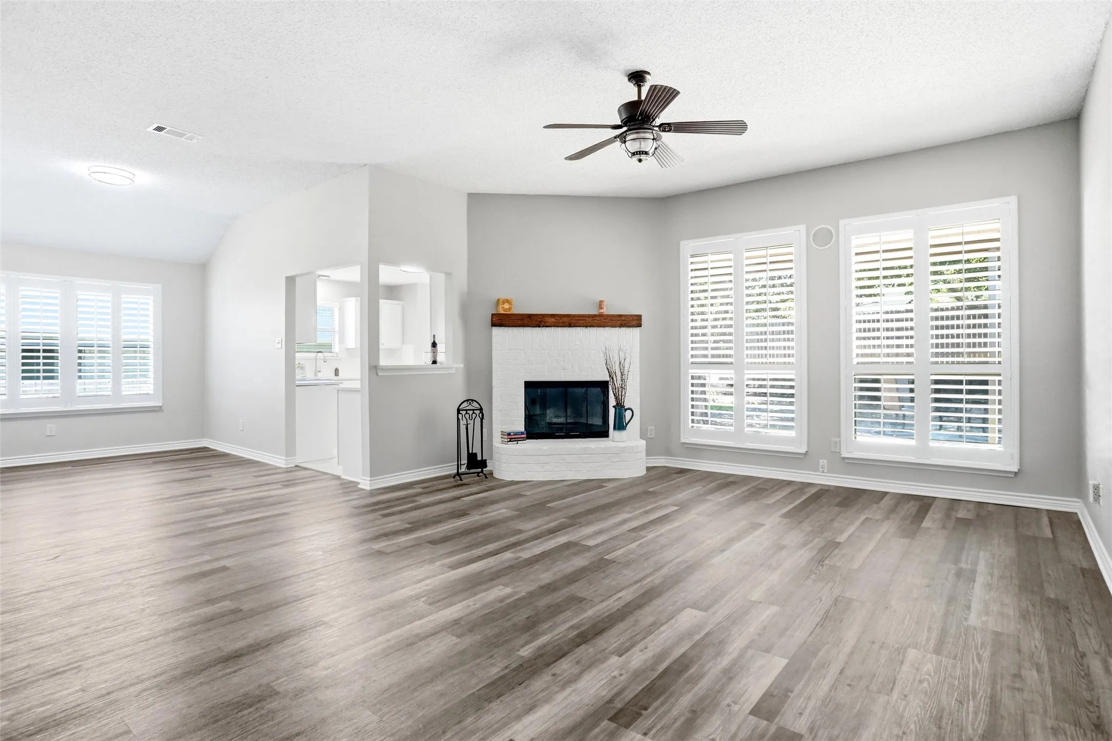 Vaulted ceiling, wood-finish flooring, white brick fireplace with wood mantel, multiple windows with white plantation shutters, and ceiling fan
