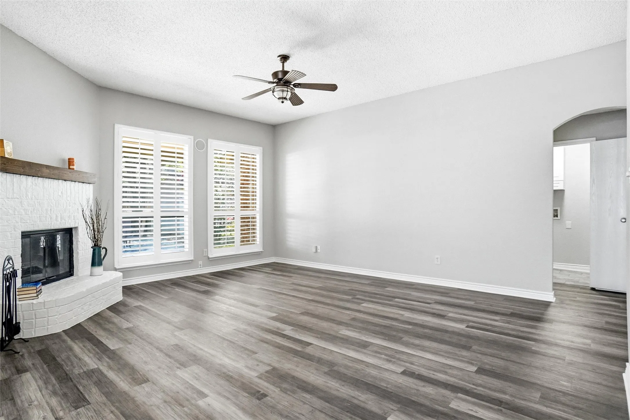 Spacious living area featuring wood-finish flooring, a white brick fireplace with a wood mantle, two windows with plantation shutters, and a ceiling fan