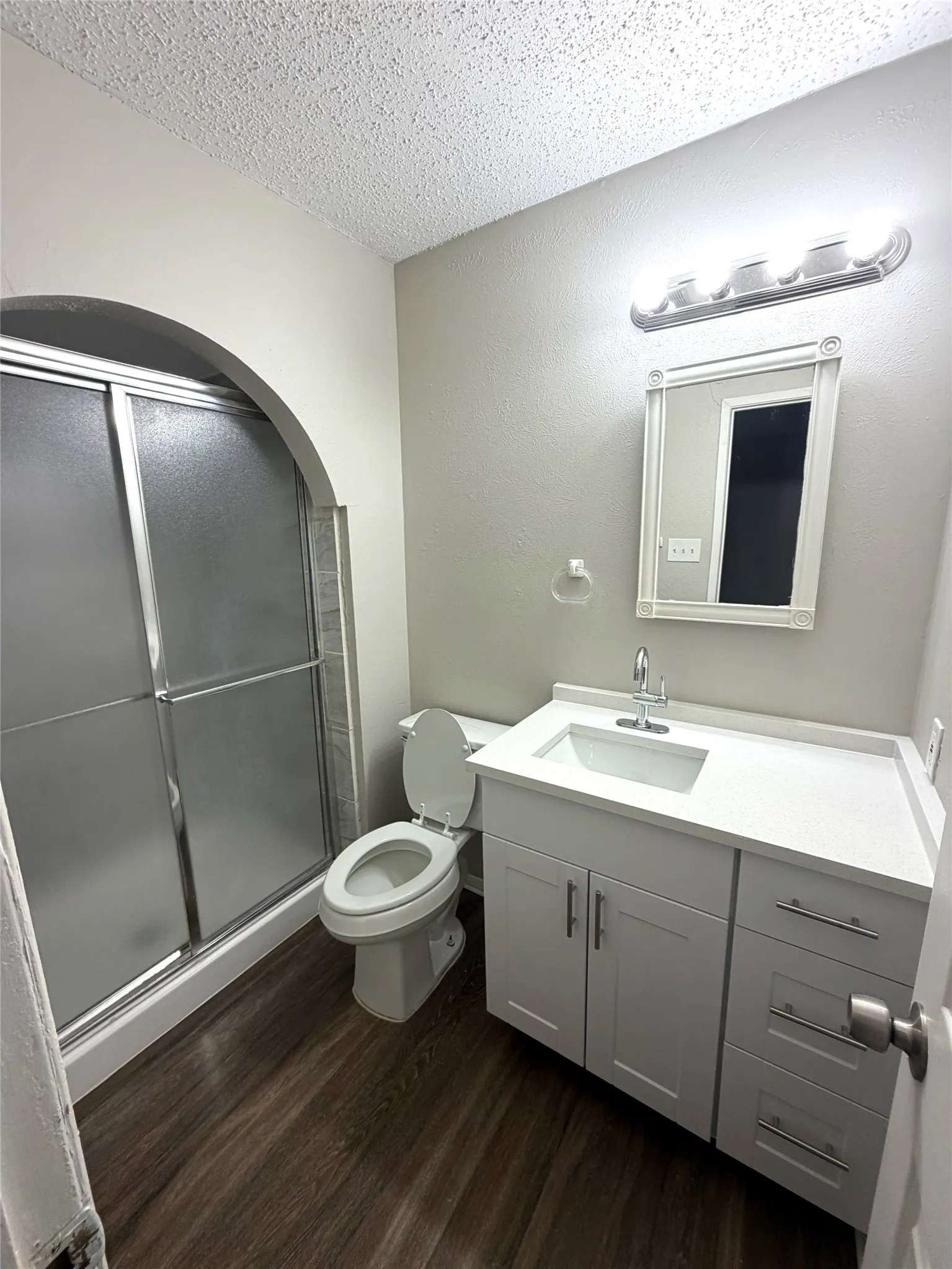 Wood-finish flooring, white vanity with integrated sink, rectangular mirror, and an arched shower enclosure
