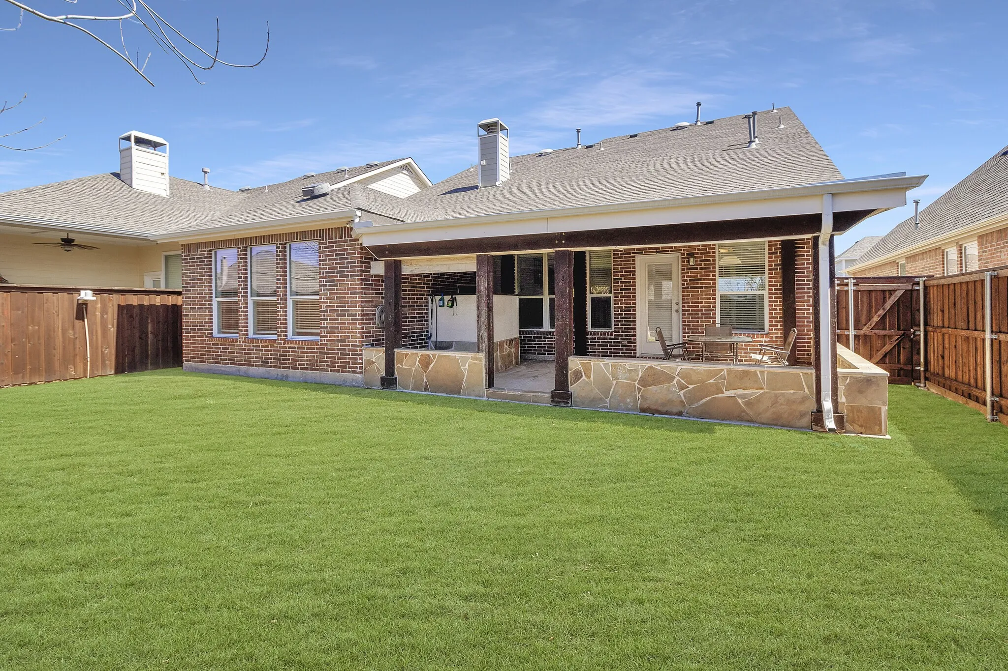 Expansive backyard with a covered patio featuring stone-clad supports, a brick exterior, and a wood privacy fence