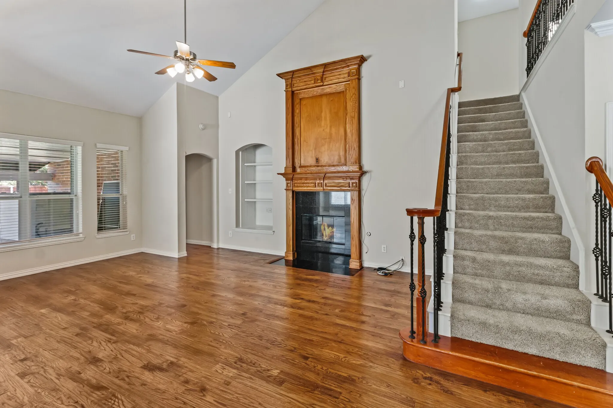 Vaulted ceiling with ceiling fan, wood-finish flooring, built-in fireplace with wood mantel, built-in shelving, carpeted staircase with wrought iron balusters