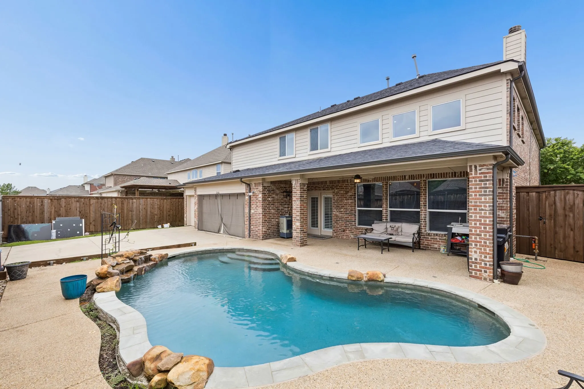 In-ground swimming pool with rock waterfall feature, exposed aggregate concrete decking, covered patio with brick columns, and wood privacy fencing
