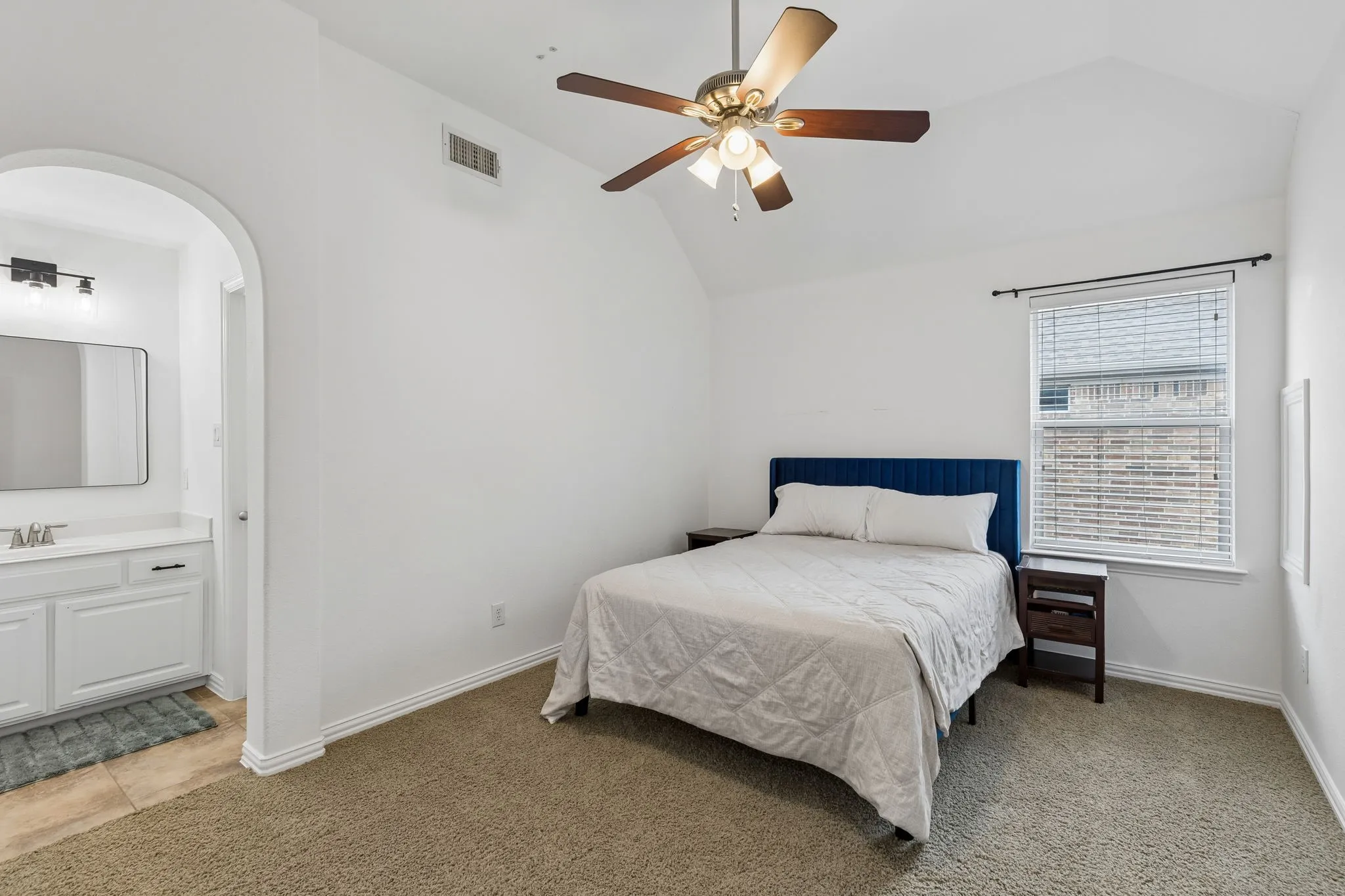 Spacious primary bedroom featuring a navy upholstered bed frame, wood-finish nightstand, abundant natural light from dual windows with blinds, ceiling fan with integrated lighting, and neutral carpeting throughout