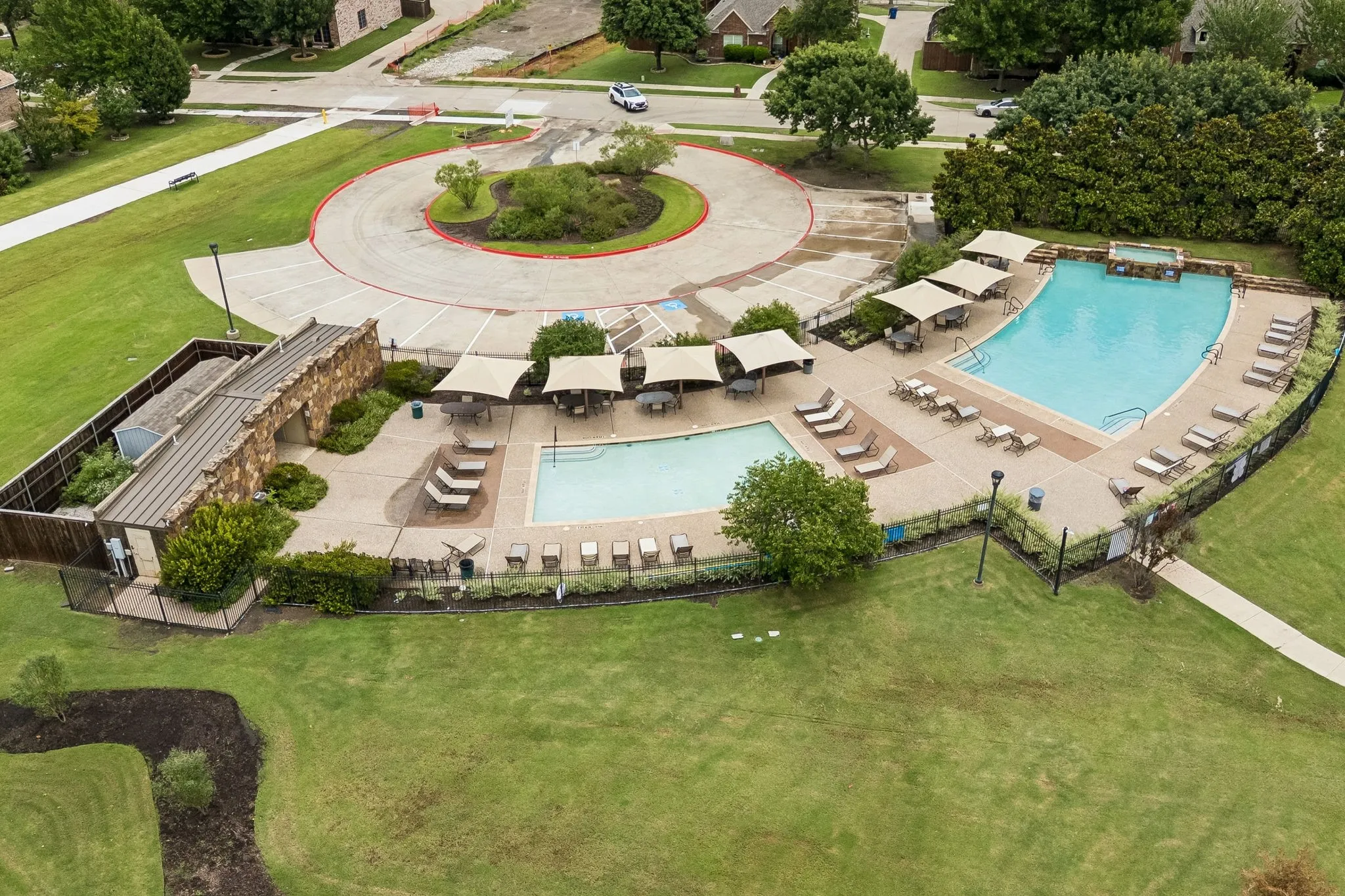 Community amenity area featuring two pools, a covered stone-clad structure, shaded seating areas, and surrounding green spaces