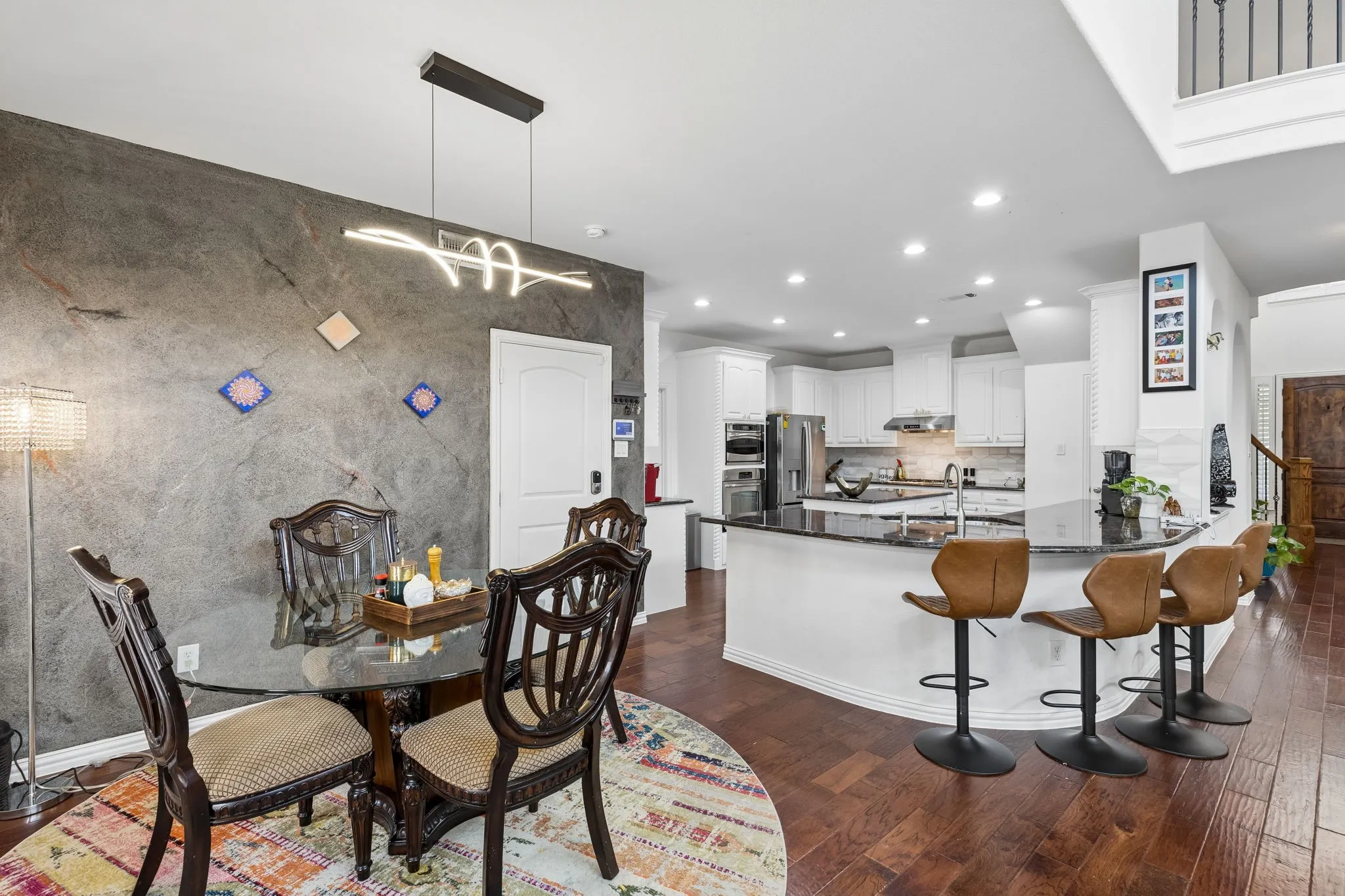 Wood-finish flooring, accent wall with decorative inlays, contemporary linear pendant light, kitchen with white cabinetry, and a dark stone countertop peninsula