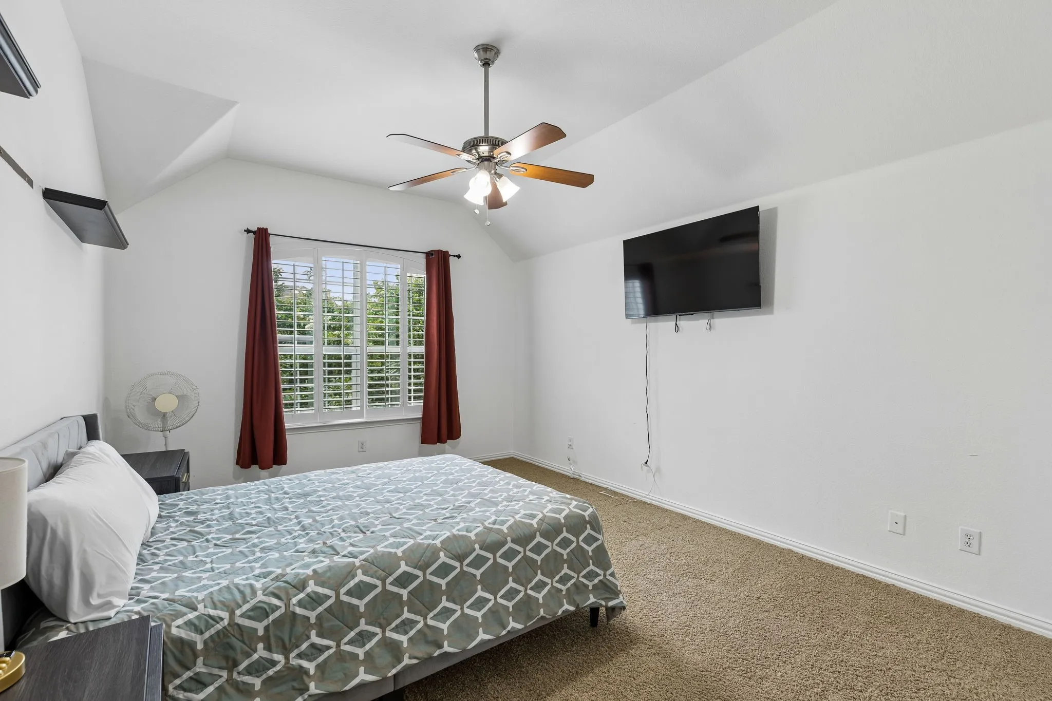 Vaulted ceiling with a ceiling fan, window with plantation shutters, wall-mounted shelving, and carpet flooring