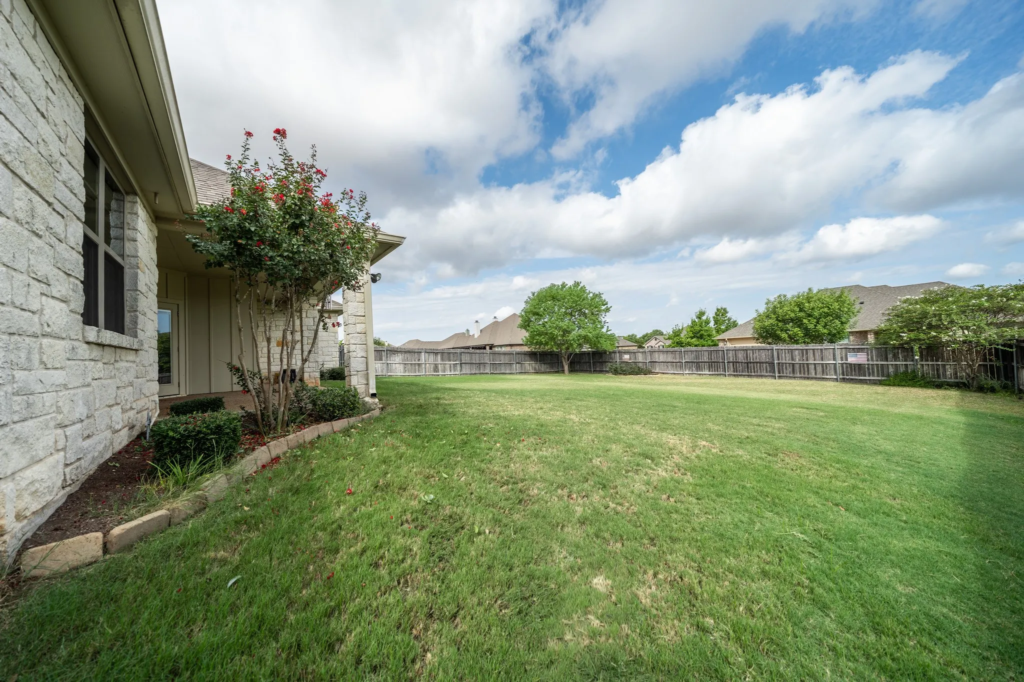 Expansive lawn area with a wood privacy fence, stone exterior, and landscaped garden beds